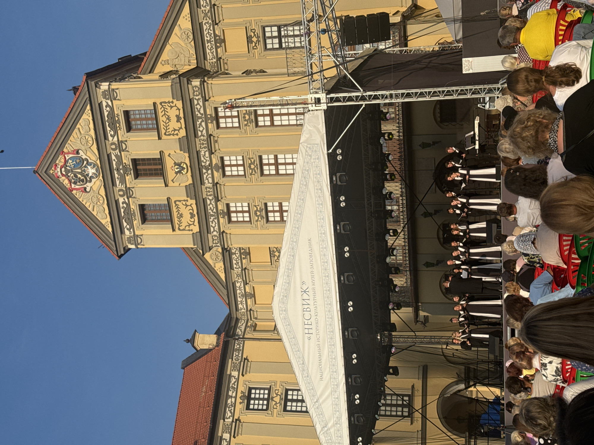 An outdoor concert is taking place in front of the Nesvizh Castle, a historic cultural landmark in Nesvizh, Belarus. A choir of approximately 12 individuals, dressed in black robes with white accents, stands on a stage, performing. A conductor in a black suit faces the choir. To the right of the choir, a keyboard player is visible. The stage is covered by a white tent with Cyrillic text that reads "НЕСВІЖ" and "НАЦЫЯНАЛЬНЫ ІСТОРЫКА-КУЛЬТУРНЫ МУЗЕЙ-ЗАПОВЕДНІК" (Nesvizh National Historical and Cultural Museum-Reserve). The audience, seated in rows of red and green chairs, faces the stage. The Nesvizh Castle, a large, ornate yellow building with a red-tiled roof, serves as the backdrop for the event. The sky above is clear and blue.