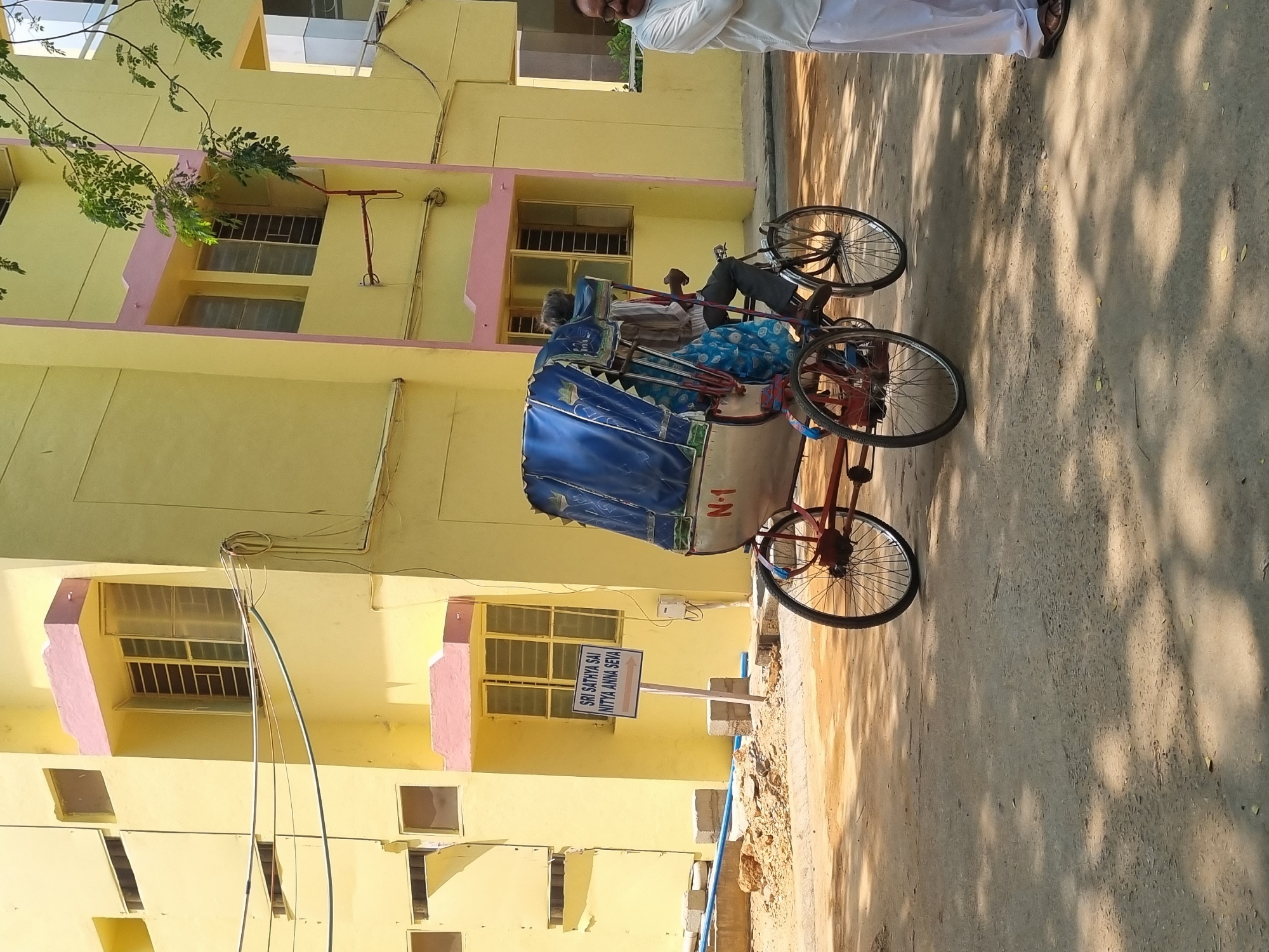 The image depicts an outdoor scene in Puttaparthi, India, specifically near a facility identified by a sign reading "SRI SATHYA SAI NITYA ANNA SEVA," indicating a location associated with the Sri Sathya Sai Central Trust, likely providing daily food service.
The scene is dominated by a multi-story building in the background, featuring pale yellow painted walls and distinct pink-colored architectural trim around windows and as vertical bands. Multiple rectangular windows with glass panes and metal grates are visible across the building's facade. Utility lines, including black cables and a white coiled cable, are attached to the building.
In the mid-ground, a cycle rickshaw, bearing the designation "N-1" on its side, is positioned on a paved surface. The rickshaw has a white passenger carriage covered by a dark blue fabric canopy and is propelled by a bicycle mechanism. A male driver, wearing a light-colored shirt and dark trousers, is seated on the rickshaw, facing generally away from the viewer.
To the right, a second male individual stands upright, facing partially left towards the rickshaw. He is wearing a white shirt, white trousers, and brown sandals. His arms are crossed, and he wears eyeglasses and has a mustache.
The ground consists of a light grey paved surface (asphalt or concrete) with visible patches of sunlight and dappled shadows, suggesting overhead foliage. To the left, closer to the building, an unpaved area shows loose dry soil, scattered rocks, and blue pipes lying on the ground. Green foliage from a tree or bush is visible in the upper right portion of the frame, extending over the building.