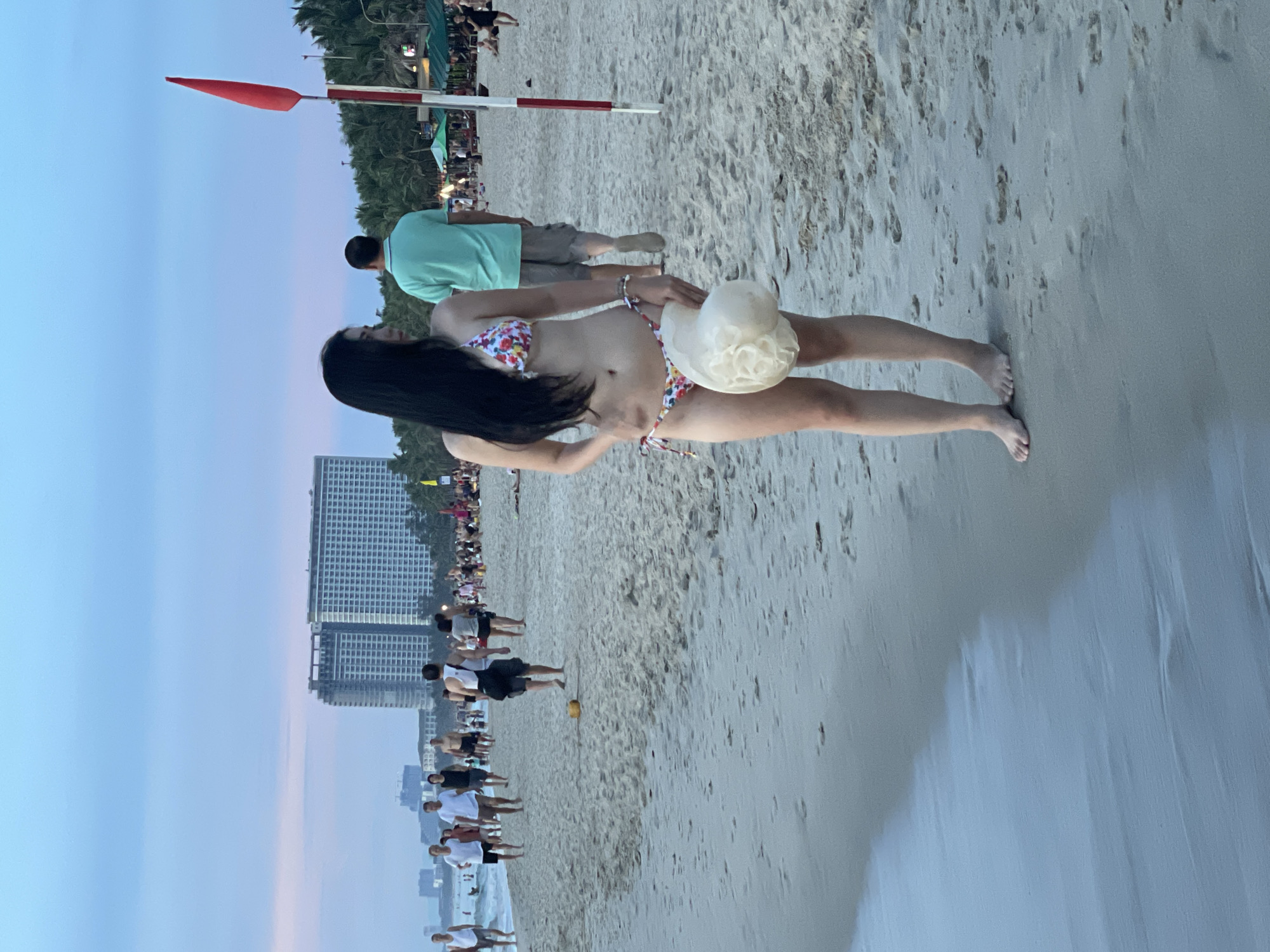 A woman in a floral bikini stands on a sandy beach, holding a light-colored hat. She is looking away from the camera to her right. Behind her, a man in a green shirt and shorts walks away from the camera. In the background, there are several people walking on the beach, two tall buildings, palm trees, and a red flag on a pole. The sky is a light blue, indicating it is either early morning or late evening. This appears to be a public beach in the city of Đà Nẵng, Vietnam, likely along the coastline.