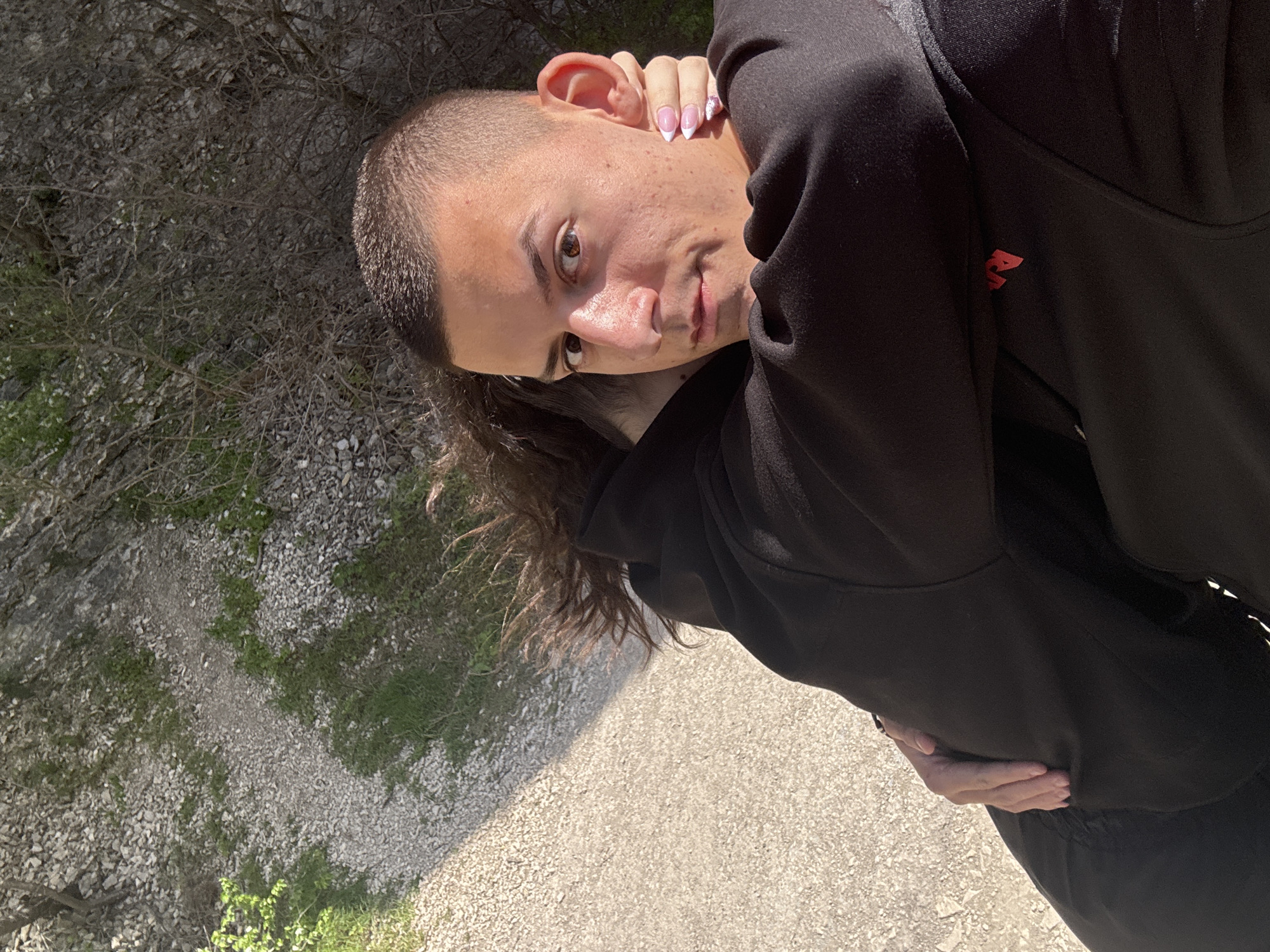 The image portrays a young individual outdoors in a natural setting, likely in Asparuhovo, Bulgaria. The person, facing the camera with a direct gaze and a slight smirk, has a distinct hairstyle featuring a buzz cut on the top of their head, contrasting with longer, curly brown hair visible along the lower and left edges of the frame. Their dark brown eyes are prominent, and their skin shows visible pores.
They are dressed in a black long-sleeved top or hoodie, which has a small red, stylized "A" logo on the left arm or shoulder area. One hand is raised towards their ear, showcasing fingernails painted in a French manicure style with a pink base, white tips, and some glitter or embellishment on one nail. The other hand rests casually on their stomach.
The background reveals an outdoor environment characterized by sparse trees and shrubs, some with bare branches and others with green foliage. The ground is covered with light-colored gravel or small stones, interspersed with patches of green grass. A clear gravel path stretches through the scene. The presence of bright sunlight, casting distinct shadows, suggests the photo was taken during midday or early afternoon under clear, sunny skies. The overall impression is a casual, sunny outdoor portrait.
