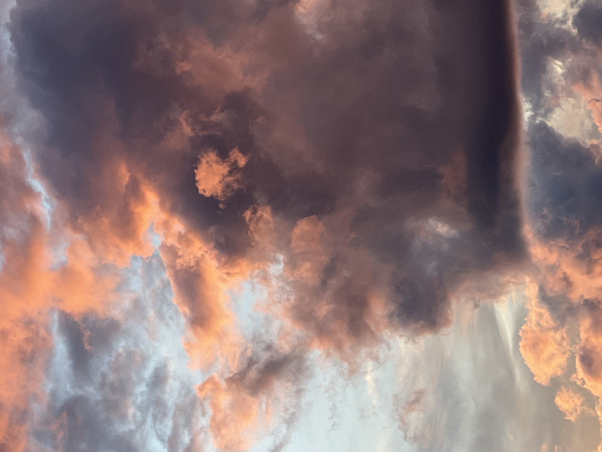 The media file captures a dramatic sky filled with clouds illuminated by the warm hues of sunset. The clouds exhibit a range of colors, from deep purples and grays to vibrant oranges and pinks, suggesting the sun is either rising or setting. The lighting creates a sense of depth and texture within the cloud formations, with some areas appearing brightly lit and others cast in shadow. The overall impression is one of natural beauty and the dynamic display of atmospheric conditions. There are no visible people, objects, text, or specific location cues beyond the general context of a sky scene. The weather appears to be cloudy, with the colors indicating either dawn or dusk.