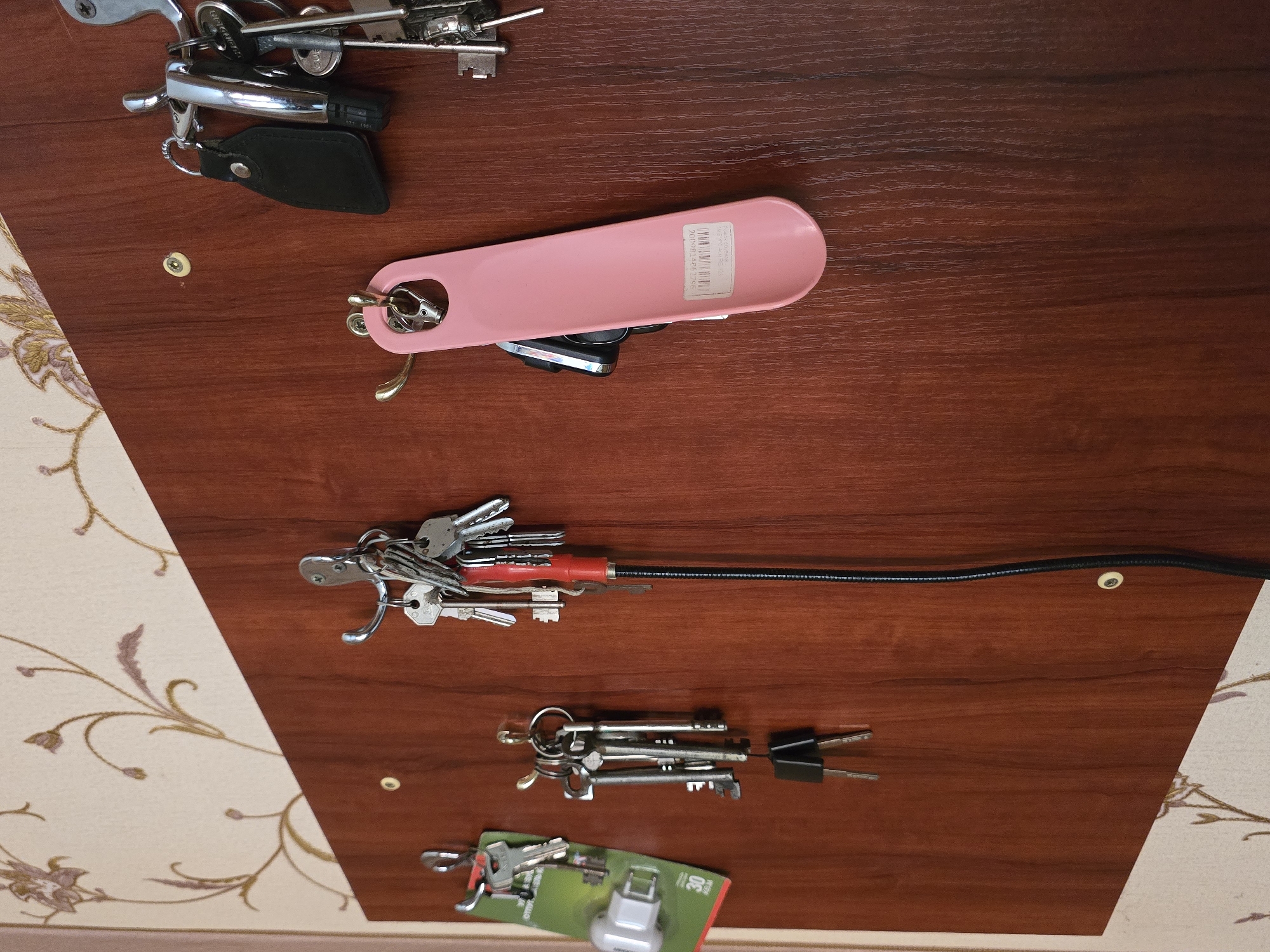 The image displays an overhead view of several sets of keys and related household items arranged on a reddish-brown wooden surface in an indoor setting. The wooden surface features a visible grain, and a section of light-colored wallpaper with a floral or vine pattern is visible on the left and bottom. Two small, light-colored fasteners are embedded in the wood, and a black cable runs across the top right.
Multiple bunches of metallic keys are present. In the upper section, a keyring holds various keys, including one attached to a black leather key fob and another to a silver remote fob. Below this, a pink, elongated object, resembling a shoehorn, is visible with keys attached. This pink item has a white sticker displaying a barcode and numbers, including "990008331306". Further down, a large collection of keys is fastened to a red-handled, flexible black rod, which appears to be a drain cleaning tool. Near the bottom, a fourth set of keys is seen alongside a small lock in green and white retail packaging, where the word "LOCK" and numbers like "100" are discernible. No people are present, and no activity is occurring. The scene is located in Nova Kakhovka, Ukraine.