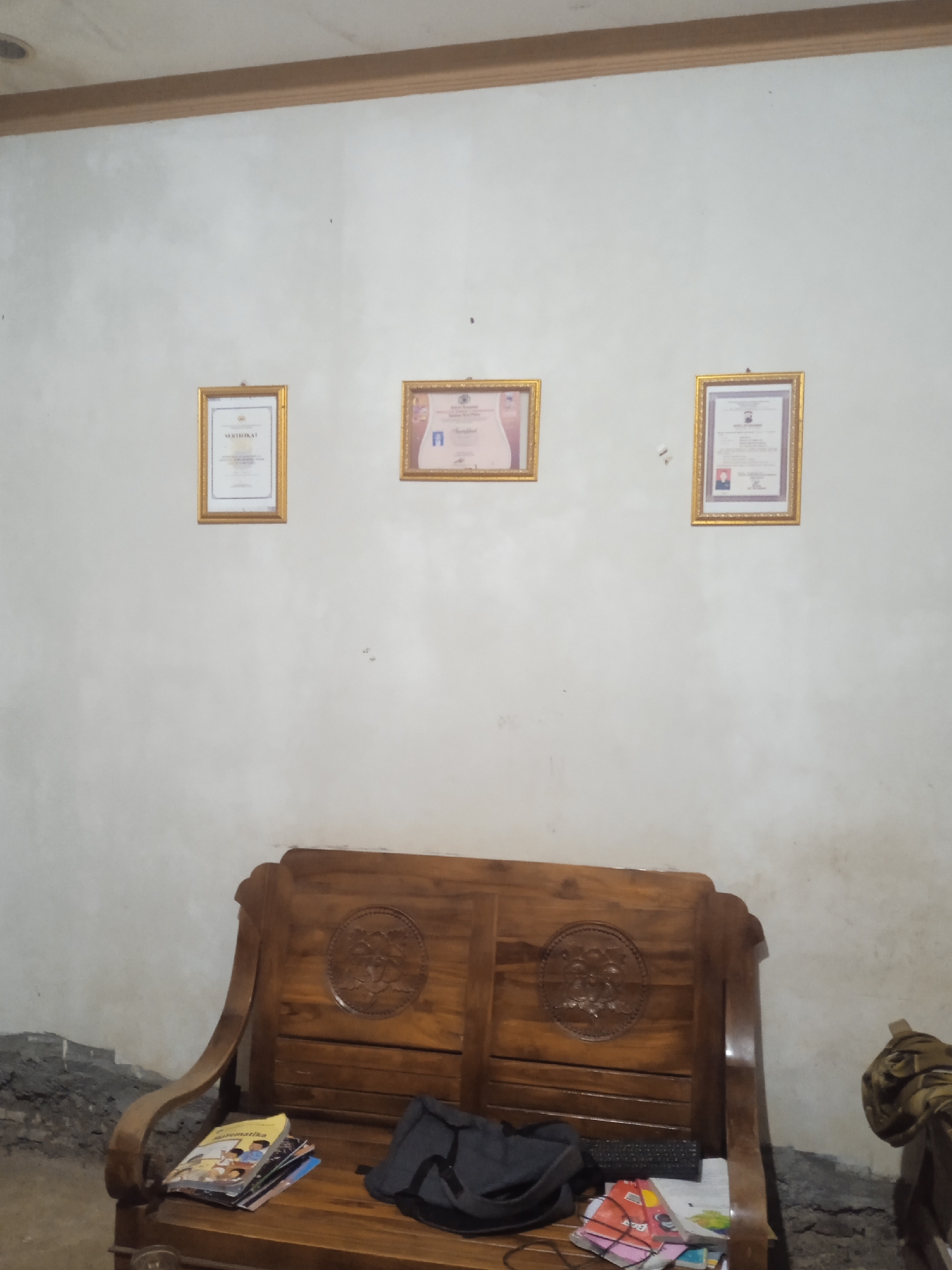 The image shows a wooden bench against a white wall in what appears to be a room in Jatisrono, Indonesia. Three framed certificates are hung on the wall above the bench. The certificates are all rectangular and appear to be gold-framed. The leftmost certificate has text that reads "SERTIFIKAT". The middle certificate has a photo of a person. The rightmost certificate also has text and a photo. On the bench, there are several books, including one with the title "Matematika" visible, and a black bag. The floor is a rough, uneven surface. The lighting is dim, casting shadows on the wall and bench.