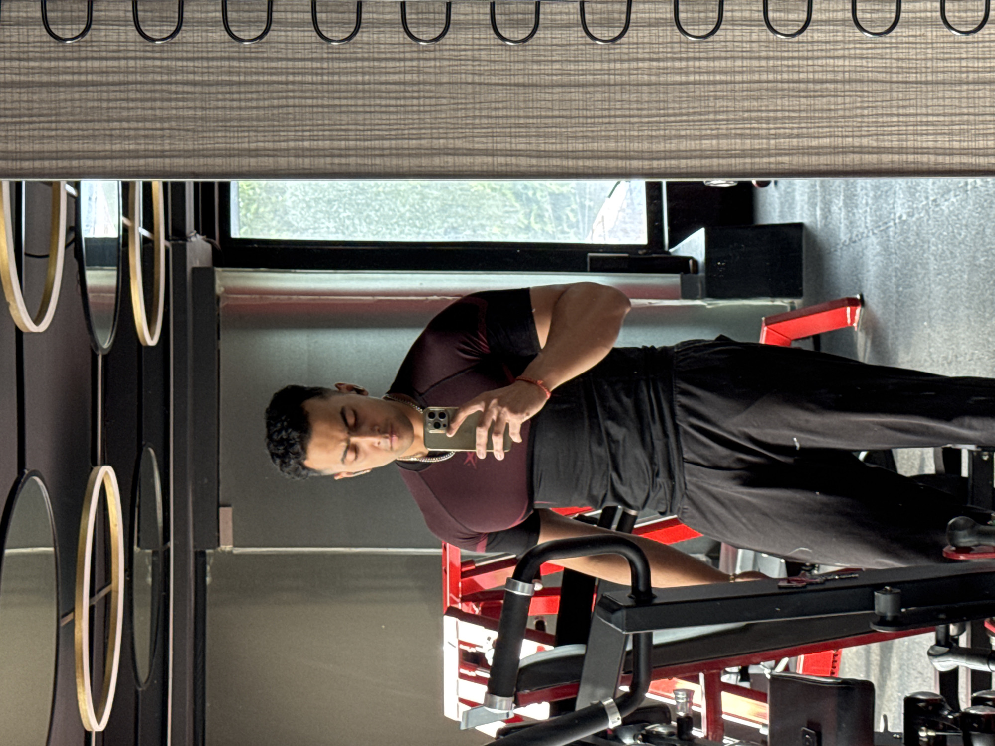 A young South Asian man, appearing to be in his early to mid-twenties, stands in a gym, taking a mirror selfie. He is wearing a two-toned athletic shirt with a maroon upper section and a black lower section, black sweatpants, and white wireless earbuds. He also has a silver chain around his neck and a red thread bracelet on his right wrist. He is holding a smartphone in his right hand, angled to capture his reflection.
The gym environment features a weight machine in the foreground on the left, with red accents visible on its structure. The background is slightly blurred, showing gym equipment and a large mirror. Above, circular light fixtures with gold accents are visible on the ceiling. To the right of the man, a wooden-patterned panel with U-shaped hooks is mounted on the wall. Through a window in the background, greenery and daylight are apparent, suggesting the gym is not underground. The overall scene depicts a casual workout session at a modern fitness facility. The image is consistent with a gym setting in a metropolitan area like Kolkata, India, due to the architecture and common gym aesthetics found in such cities.