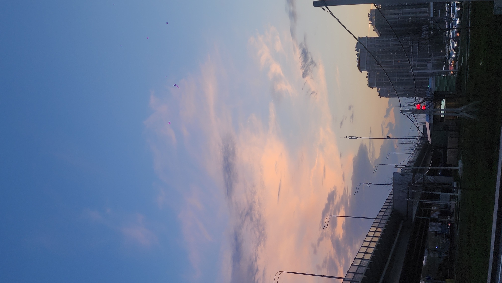 The image shows a sunset scene in Moscow, Russia. In the foreground, there is a highway overpass with streetlights. A building is on the right side. A traffic light is visible. The sky is mostly blue with pink and purple-tinged clouds. There appear to be some purple dots in the sky, likely balloons or similar objects.