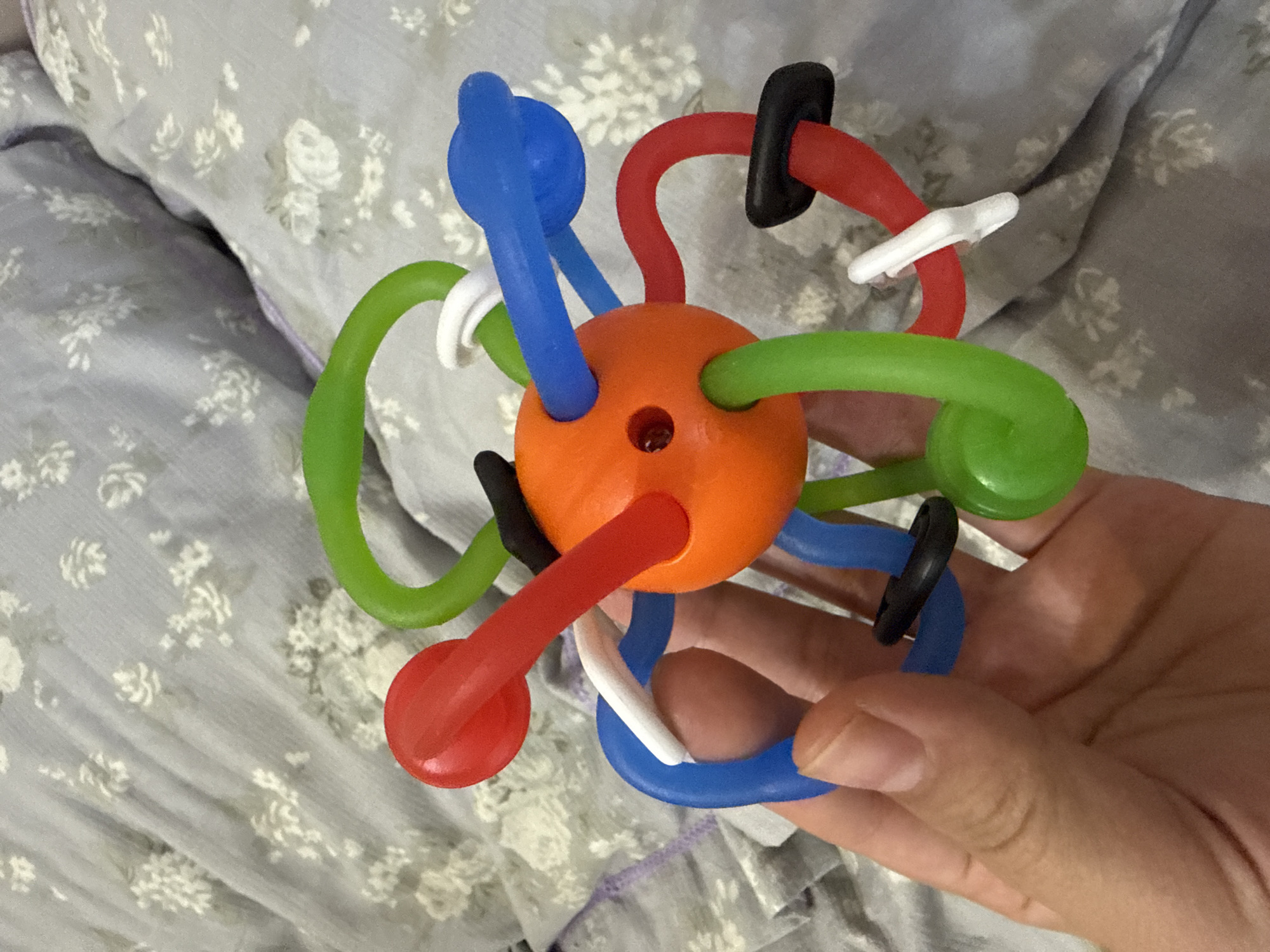 An orange, spherical baby toy with flexible, colorful arms is held in a person's left hand. The toy has red, green, and blue arms, each ending in a round knob. Some of the arms also have black or white clips attached. The toy is positioned on a light gray blanket with a faint white floral pattern. The background is out of focus, showing more of the same patterned blanket.