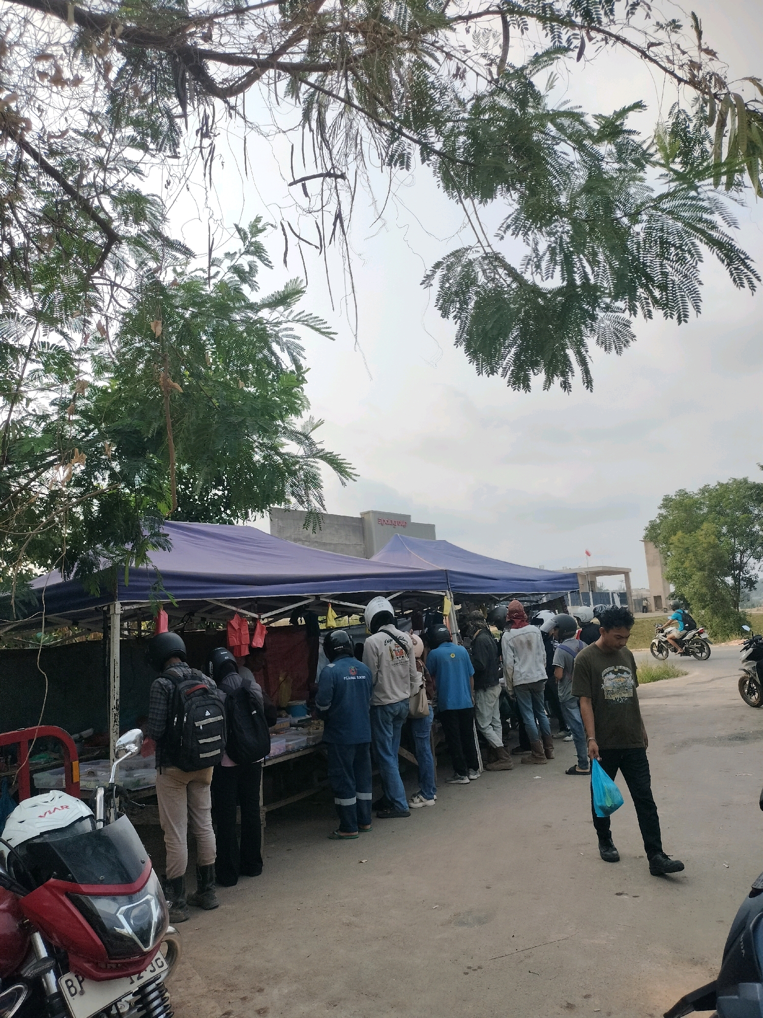 This is a medium-shot, outdoor, eye-level photograph of a street scene in Batam, Indonesia. In the foreground, a red motorcycle is parked. Behind it, several people are gathered around a stall with a blue tarp. They are queuing and appear to be purchasing items. The stall is set up along a paved road. In the background, a building with signage is visible, and a person is riding a motorcycle on the road. The sky is overcast.