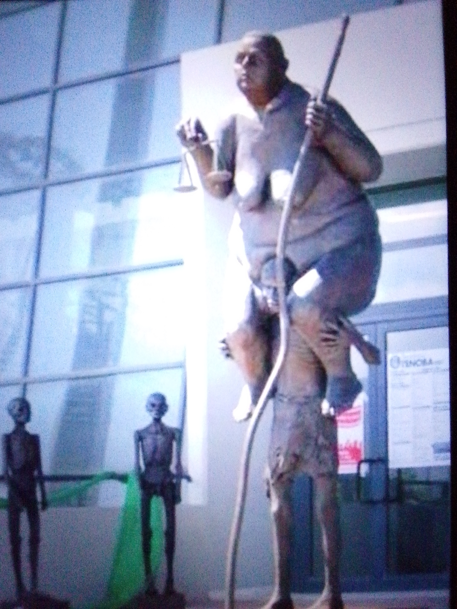 The image shows a blurred, low-resolution statue of a man standing on top of another statue, which is likely also a man. The top statue is holding a scale in its right hand and a rod in its left. The bottom statue is in a crouching or kneeling position. In the background, there are two slender, dark figures that appear to be statues or mannequins, one in the left middle ground and another to its right. A vibrant green fabric is draped behind the two figures. Behind the statues, a modern building with large, gridded windows is visible. There are no discernible interactions between the statues or figures. The lighting suggests it is daytime.
