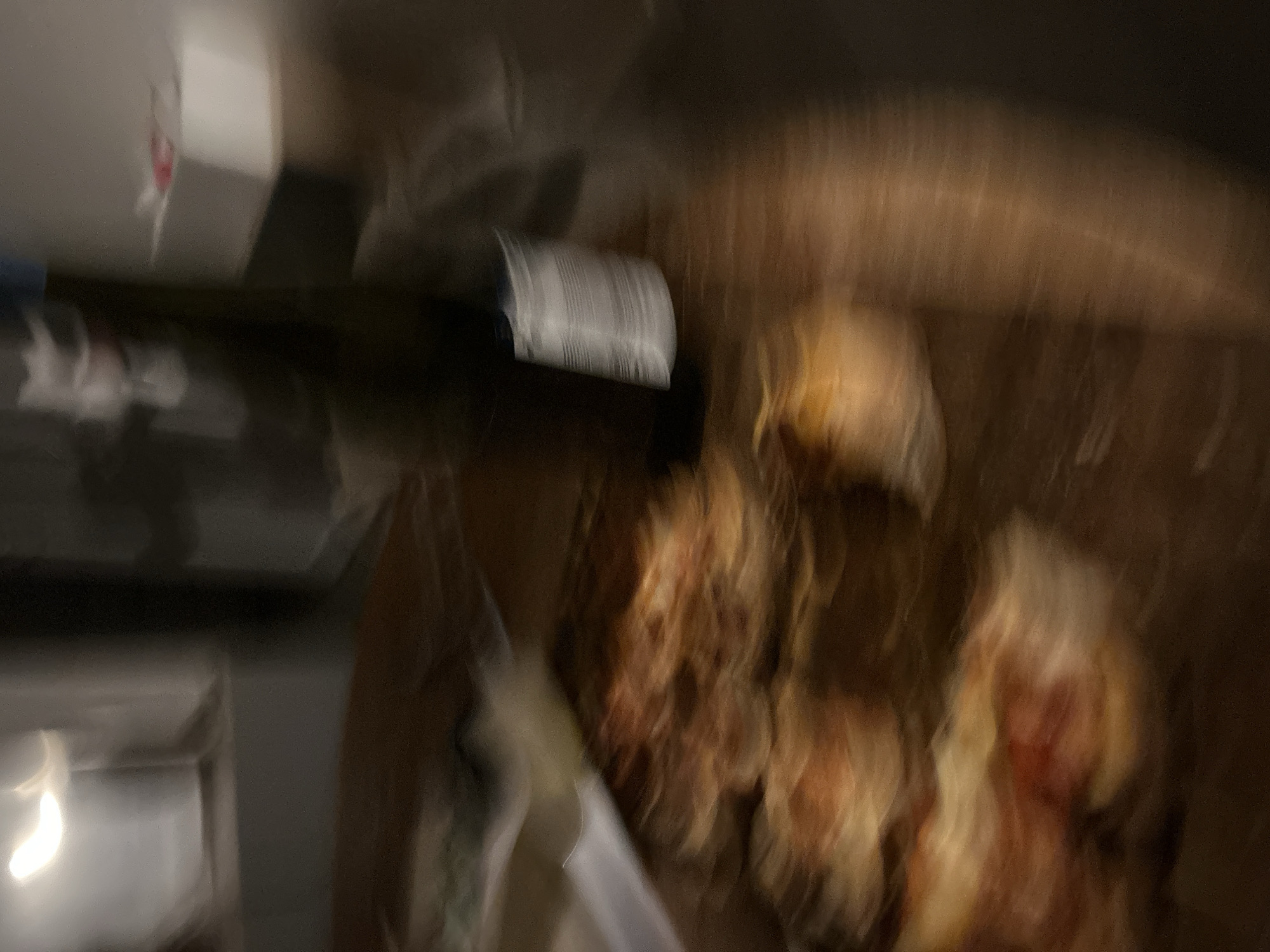 The image is heavily blurred, depicting an indoor scene captured in low light with significant motion.
**Subjects:**
The primary subjects are several irregularly shaped, light golden-brown objects, likely food items such as pastries, baked goods, or fried products, with a textured, possibly crusty or flaky surface. They appear clustered together, perhaps in a container or on a surface. To the left, there is a dark, elongated object resembling a bottle, featuring a distinct white label with illegible text or a barcode. Further left and partially visible is a white, rectangular object, possibly a box or another container, with a faint red mark on it.
**Scene and Setting:**
The setting appears to be an indoor environment, possibly a kitchen counter or a table, indicated by the presence of food and a bottle. The background is dark and indistinct due to the blur, suggesting a dark surface or wall.
**Activity or Event:**
Due to the intense blur, no specific activity or event can be definitively determined. The image appears to be a fleeting snapshot of items placed on a surface, possibly in the process of being prepared, served, or unpacked. The motion blur suggests either camera movement or movement of the subjects during exposure.
**Notable Details:**
The low light conditions suggest the image was taken in the evening or at night. There are no specific location cues visible within the image itself beyond it being an indoor space. No people are visible. The text on the bottle label is too blurry to be read.