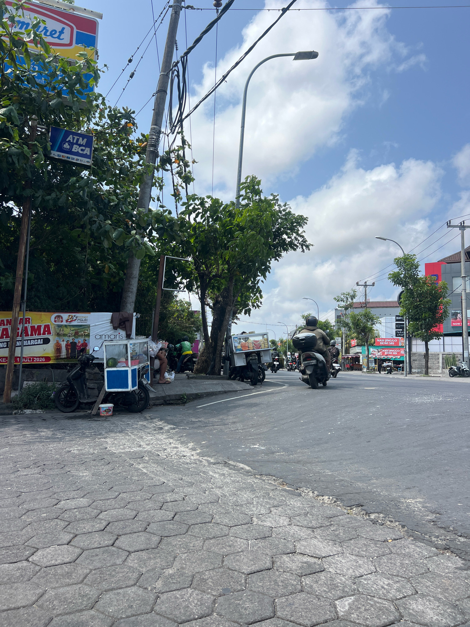 The image depicts a street scene in Kuta, Indonesia, on a bright, partly cloudy day. The foreground shows a street paved with hexagonal grey cobblestones, transitioning to asphalt. A small food cart with a blue and white facade, containing what appears to be fruit, is parked beside a black motorcycle. A person is seated on a curb near the cart.
Further down the street, several motorcycles are visible, some with riders. One motorcycle in the mid-ground has a rider wearing a helmet and a dark jacket. Another motorcycle in the background also has a rider. To the left, a large banner with images and text, including "SANTAP AR....AGAMA" and "amaris," is displayed. Above the banner, a sign for "ATM BCA" is mounted on a utility pole, which is covered with electrical wires and cables. A street lamp is also visible on this pole.
On the right side of the street, buildings can be seen, one with a red and white facade and another with a darker exterior. Signs are present on these buildings, including one that reads "WARUNG NASI JINGGO DEWI SRI." Trees line the street, some with full green foliage and others with sparser leaves. The overall impression is of a busy commercial street with pedestrian and vehicular activity.