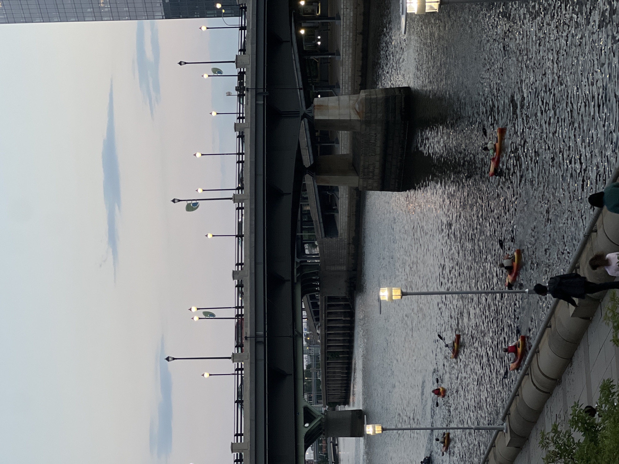 The image depicts a twilight scene on the Schuylkill River in Philadelphia, Pennsylvania. Several kayakers are paddling on the water, with some in red and yellow kayaks. A bridge spans the river, with cars visible on its surface. The riverbank features a walkway lined with lampposts. In the background, a tall building suggests an urban environment. The sky is a soft gradient of gray and pale blue, with wisps of clouds. The water reflects the dim light, creating a shimmering surface.