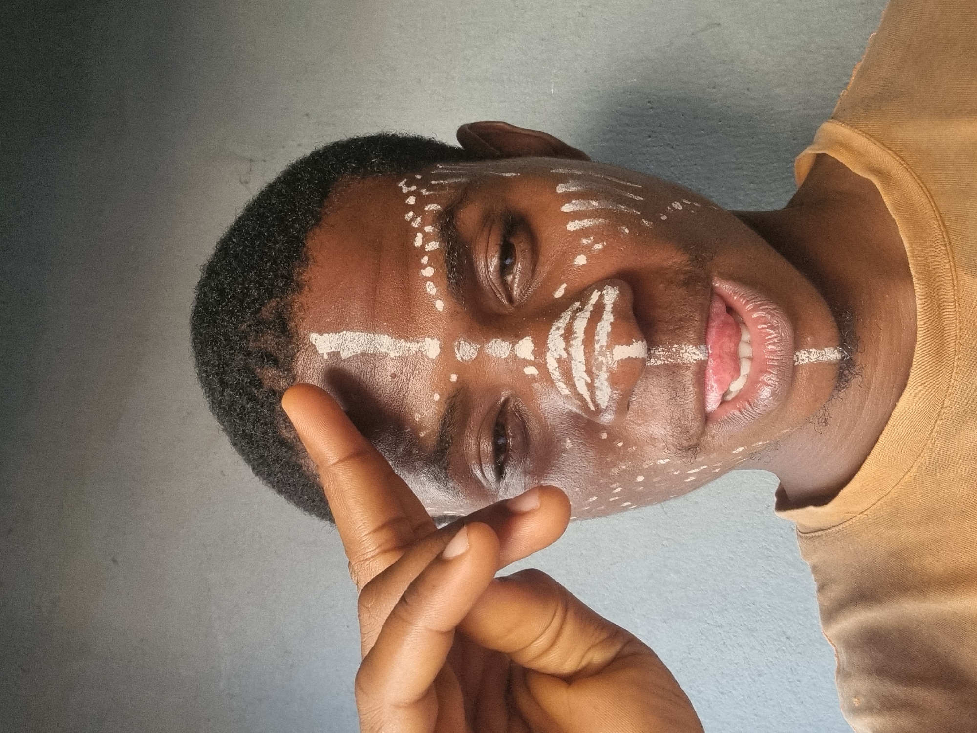 A close-up, tilted, medium shot shows a dark-skinned man with white markings on his face, and holding his hand up in front of him, with his index finger extended upward. The man's hair is dark and short. The markings consist of dots and stripes on his forehead, cheeks, and chin. There is a vertical stripe on his nose with curved stripes on the nose itself. His lips are parted slightly, and a portion of his tongue is visible. The man is wearing a golden-yellow shirt. The background is a plain, light-blue-grey wall. The location and city are unknown.