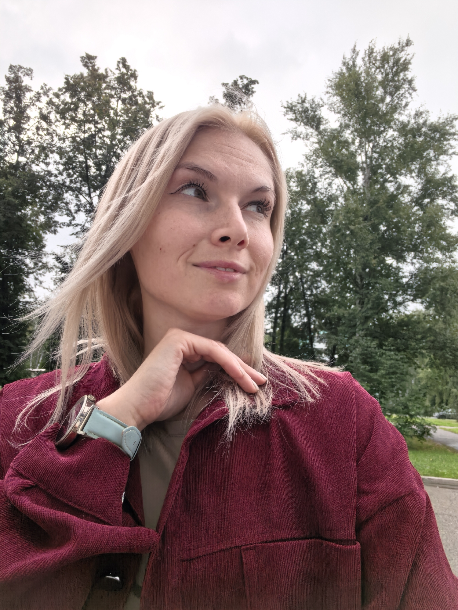 A medium shot captures a blonde woman in a maroon corduroy jacket, with her right hand touching her chin and neck. She is looking off to her right with a slight smile. Her hair is blowing in the wind. Behind her, trees and a cloudy sky are visible. The location is Sterlitamak, Russia.