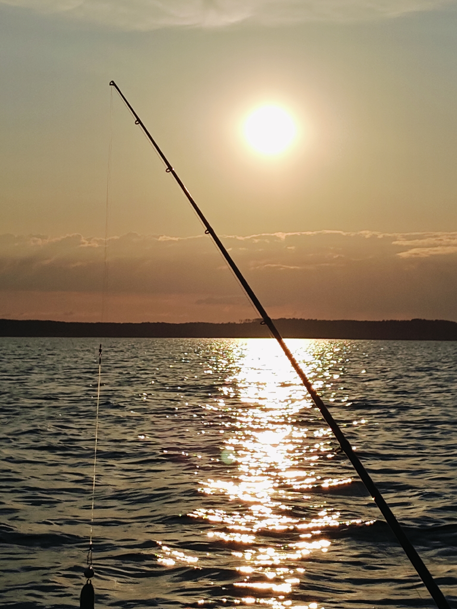 This is a photograph of a fishing rod silhouetted against a sunset over the Kama River in Chaykovsky, Russia. The sun is a bright, diffused orb in the upper right portion of the sky, casting a shimmering reflection on the water. The fishing line extends from the rod into the water, with a lure visible near the surface. The opposite bank of the river is lined with trees and appears dark against the colorful sky. The water is rippled, indicating a slight breeze. The scene evokes a sense of peacefulness and solitude.