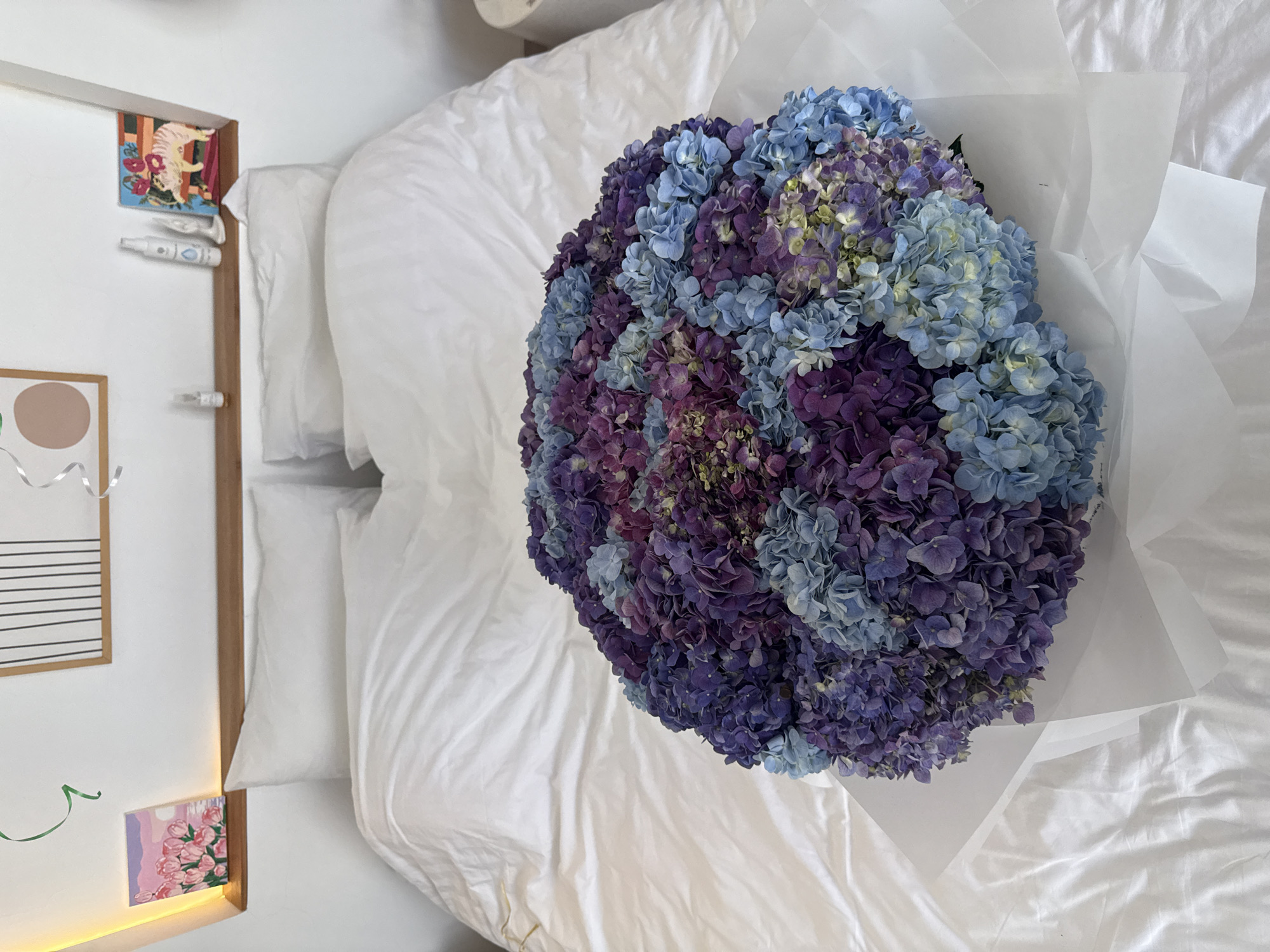 The image depicts a high-angle shot of a bouquet of flowers resting on a white bed. The bouquet primarily consists of blue and purple hydrangeas, wrapped in white paper. The bed is made with white sheets and pillows. To the left of the bed, part of a white wall and a wooden frame with decorations (including a drawing and a postcard) are visible. The location is not discernible from the visual information provided and the location name is not to be included.