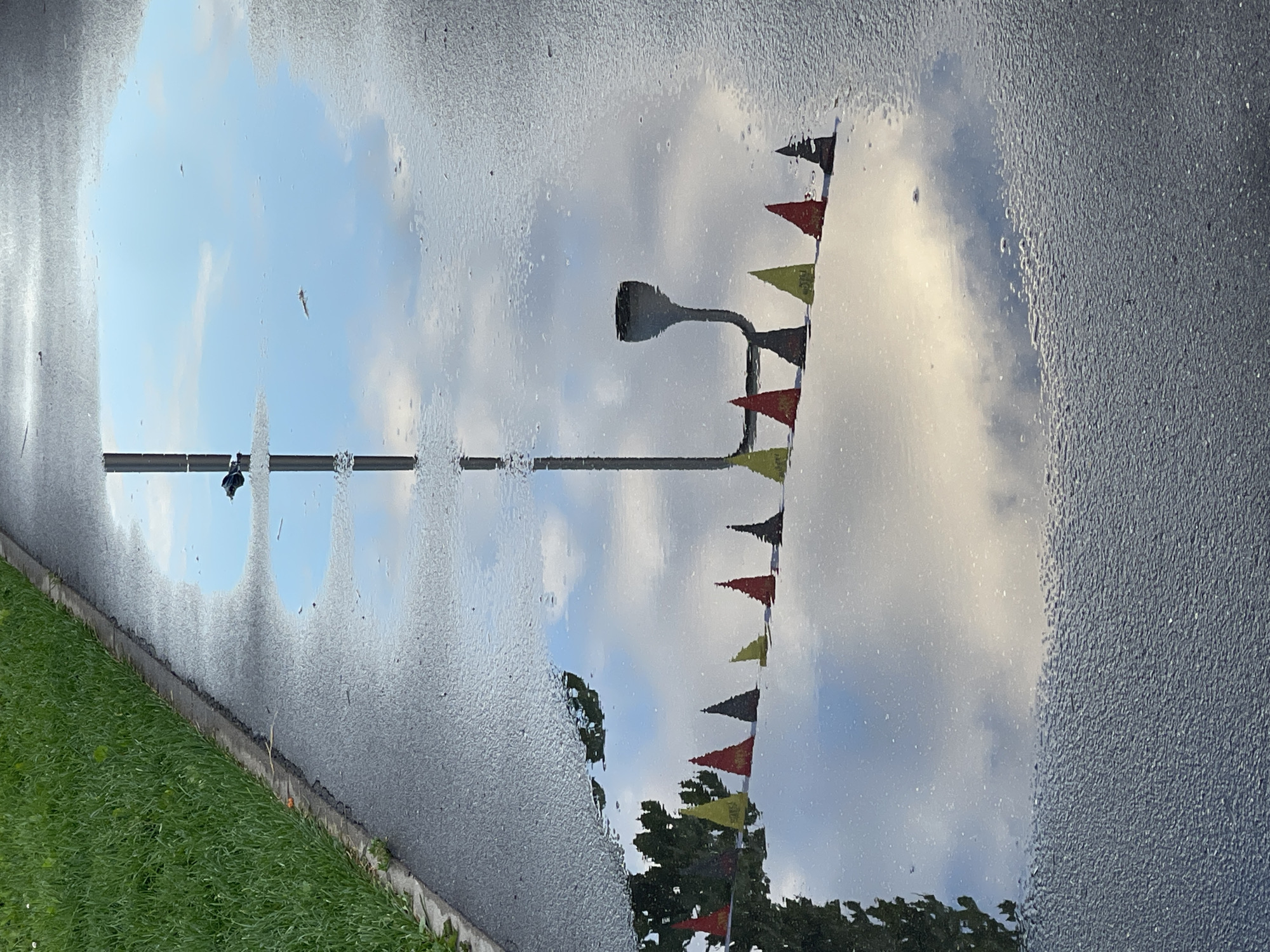 This is a photograph of a puddle on an asphalt surface next to a patch of green grass. The puddle reflects the sky, clouds, a lamppost, and a string of triangular flags in red, yellow, and dark gray. The flags are arranged in a line and appear to be part of a decoration. The scene is likely located in Khimki, Russia, given the context.