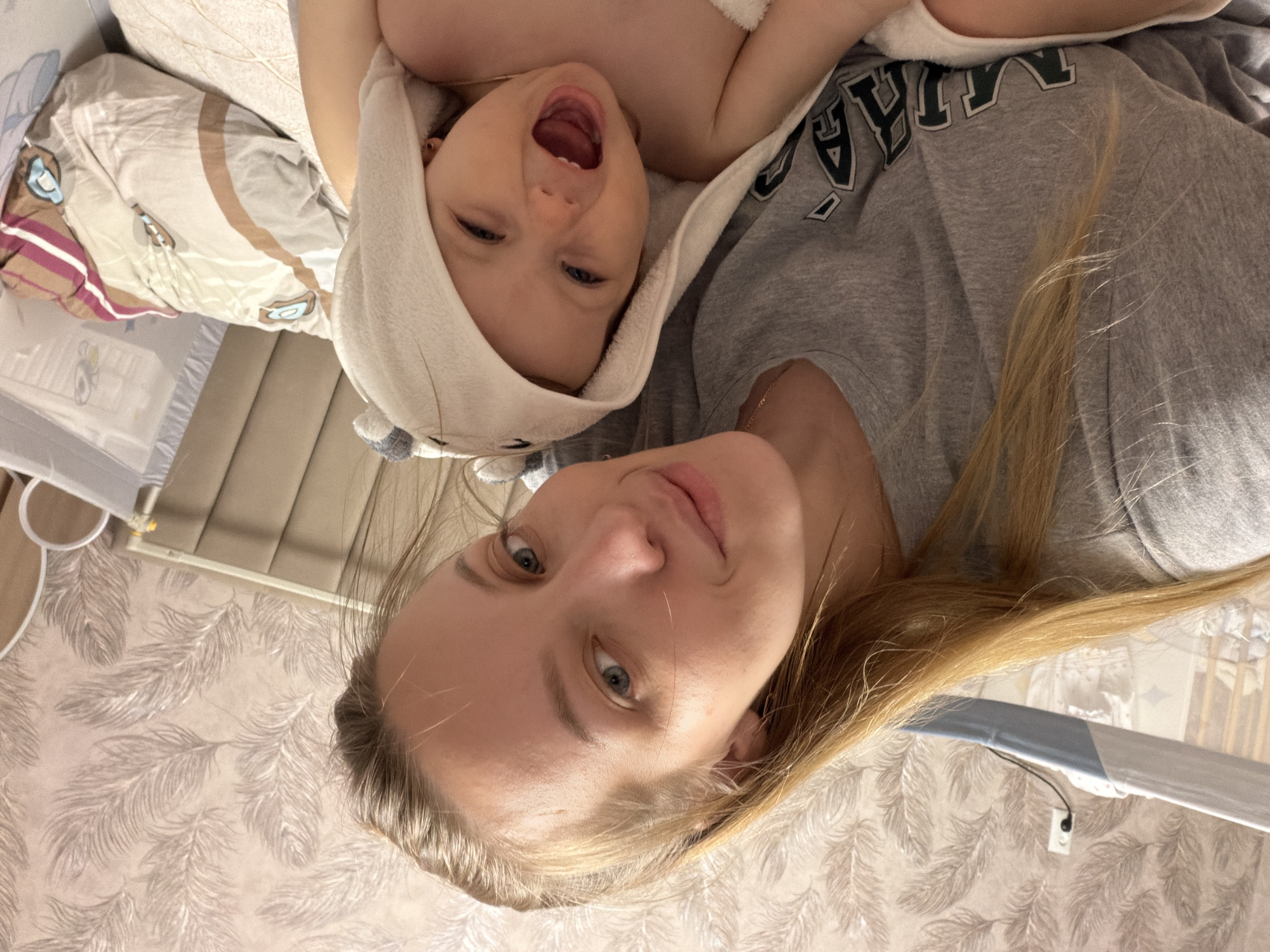 A woman and a baby are lying on a bed. The baby is wrapped in a hooded towel and is facing upwards with their mouth open. The woman is next to the baby, also facing upwards. The room appears to be a bedroom with a bedside crib partially visible. The location is unknown and there is no information about being in the city of Reshetnikovo, Russia.
