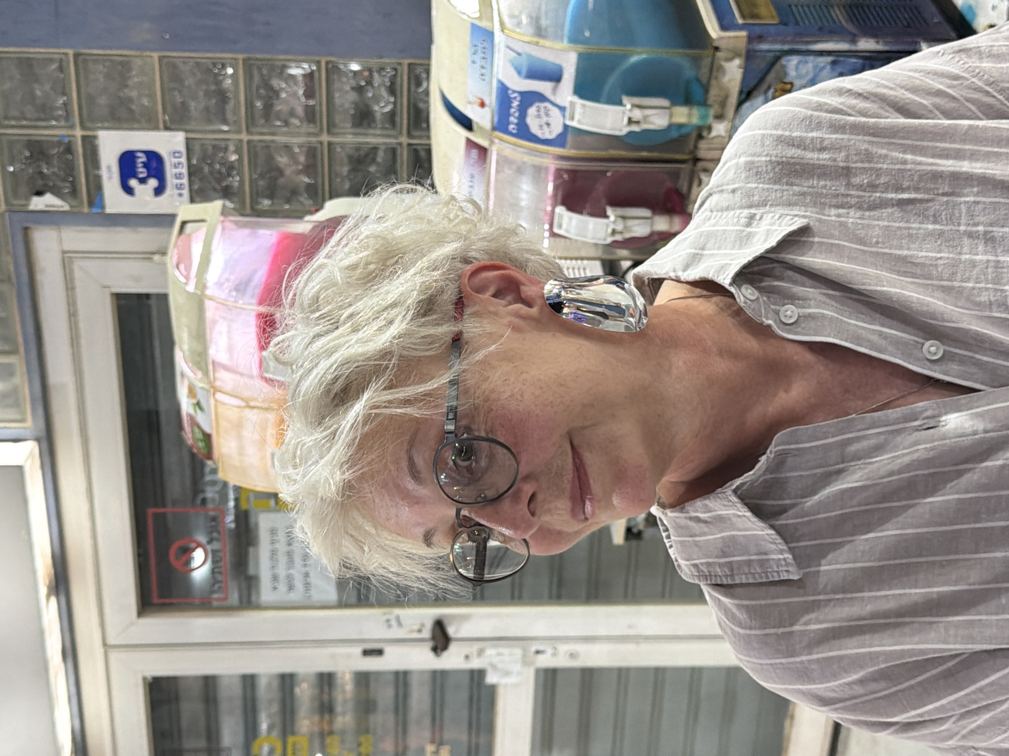 An elderly woman with white hair and glasses is standing in front of a store in Eilat, Israel. She is wearing a striped shirt and large earrings. Behind her, there are colorful slushy machines and a sign with Hebrew writing. The store appears to be a convenience store or a snack shop.