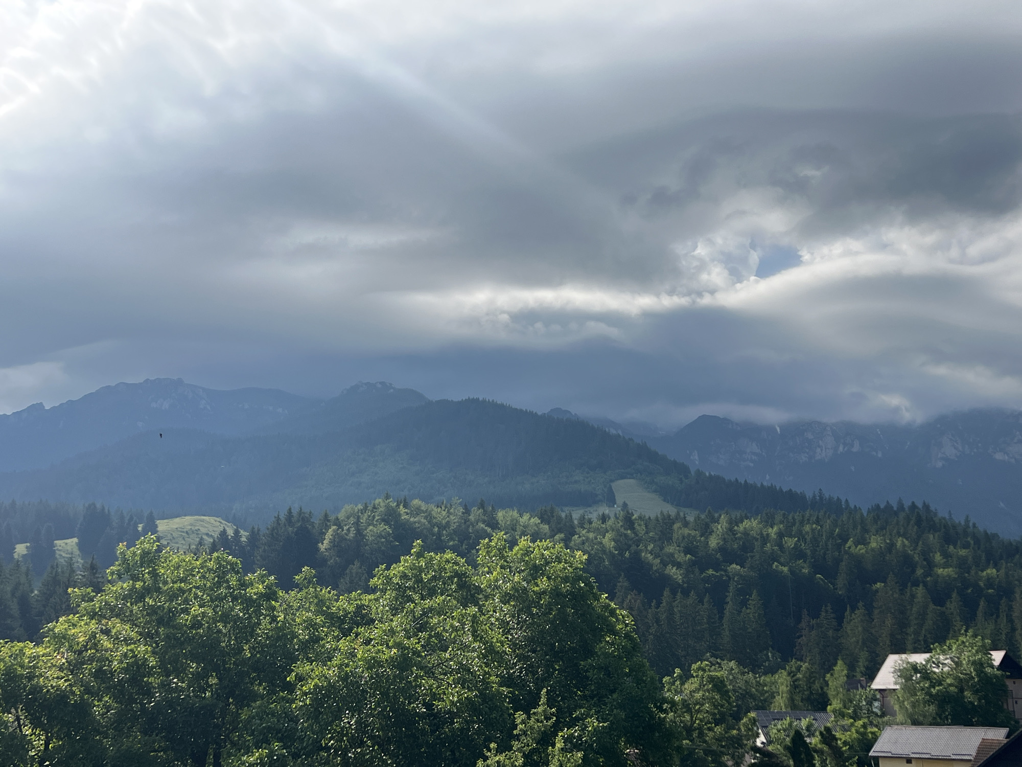 A wide-angle, eye-level shot captures a mountainous landscape partially obscured by dramatic, overcast skies. In the foreground, lush green trees dominate the lower half of the frame, with a few houses partially visible among them on the right side. The middle ground is a dense forest of evergreen trees, leading up to rolling hills and larger mountains in the background. The mountains are shrouded in a bluish haze, with rugged peaks peeking through the darker, cloud-covered sky. A sunbeam pierces through the clouds on the left, casting a bright ray across the scene. There are no visible people or animals. The overall impression is one of a serene, natural environment under a moody sky.