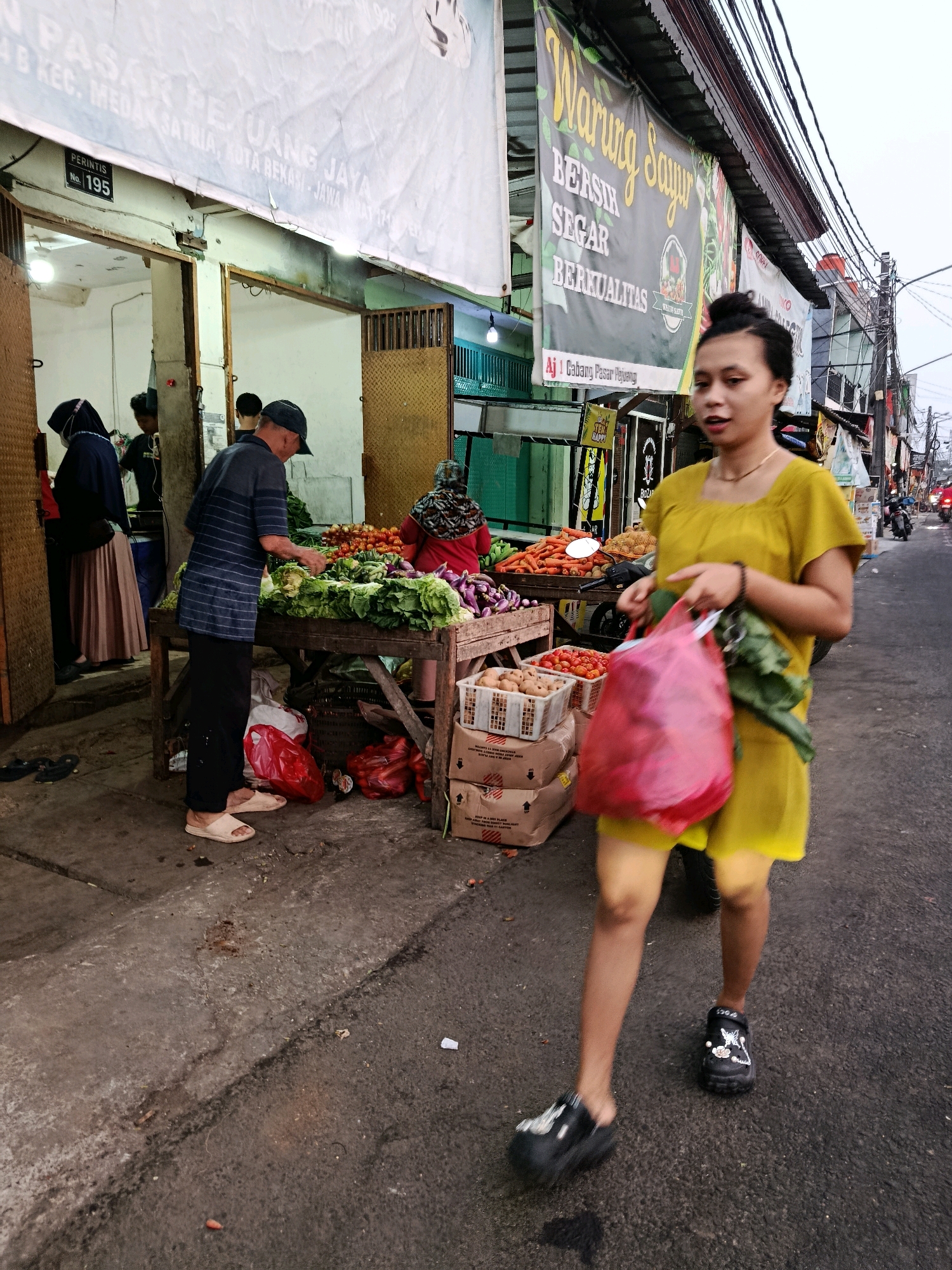 In front of a market stall in Bekasi, Indonesia, a woman wearing a yellow dress walks towards the camera, carrying a red plastic bag and some green vegetables. The market is lined with stalls selling fresh produce, including tomatoes, eggplants, and leafy greens. A man wearing a blue and white striped shirt and a cap is arranging vegetables on a table. Another person, wearing a hijab, is seen entering a shop. The street is paved and shows signs of wear and tear. Banners with text in Indonesian are visible on the stalls, indicating it is a busy marketplace.