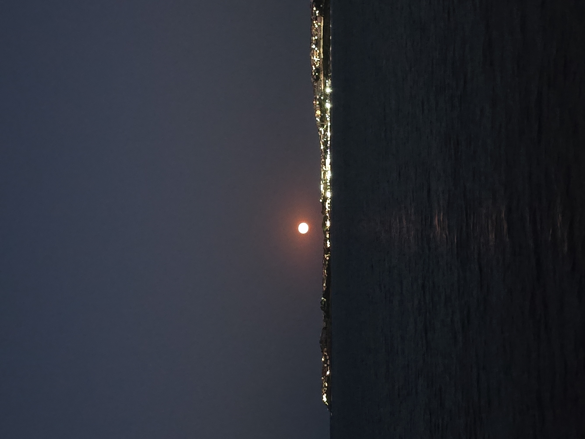 The image depicts a nighttime scene of the Chernomorets coast in Bulgaria. A large, reddish moon is visible in the dark sky, slightly to the right of the center. Below the moon, a strip of land is illuminated by numerous artificial lights, suggesting a populated area or a town. This landmass extends horizontally across the middle ground of the image, separating the sky from the dark, rippling water in the foreground. The water reflects some of the light from the moon and the distant town, creating subtle luminous streaks. The overall atmosphere is serene and somewhat ethereal, characteristic of a moonlit night over the sea.
