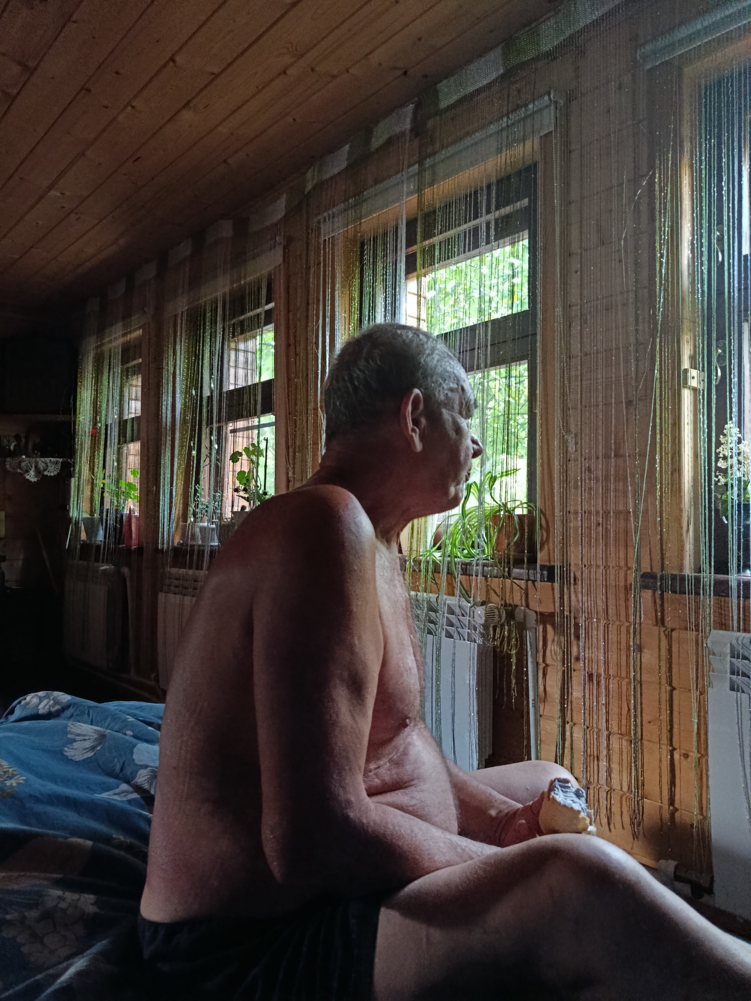 An older man, shirtless and wearing dark shorts, sits on a bed covered with a blue and white floral patterned comforter. He is looking out a window that is adorned with hanging string decorations. Sunlight streams through the window, illuminating plants on the windowsill. The room has wooden walls and a wooden ceiling, with a radiator visible below the window. The man is holding a piece of food in his hand. The scene appears to be in a rustic setting, possibly a cabin or a rural home.