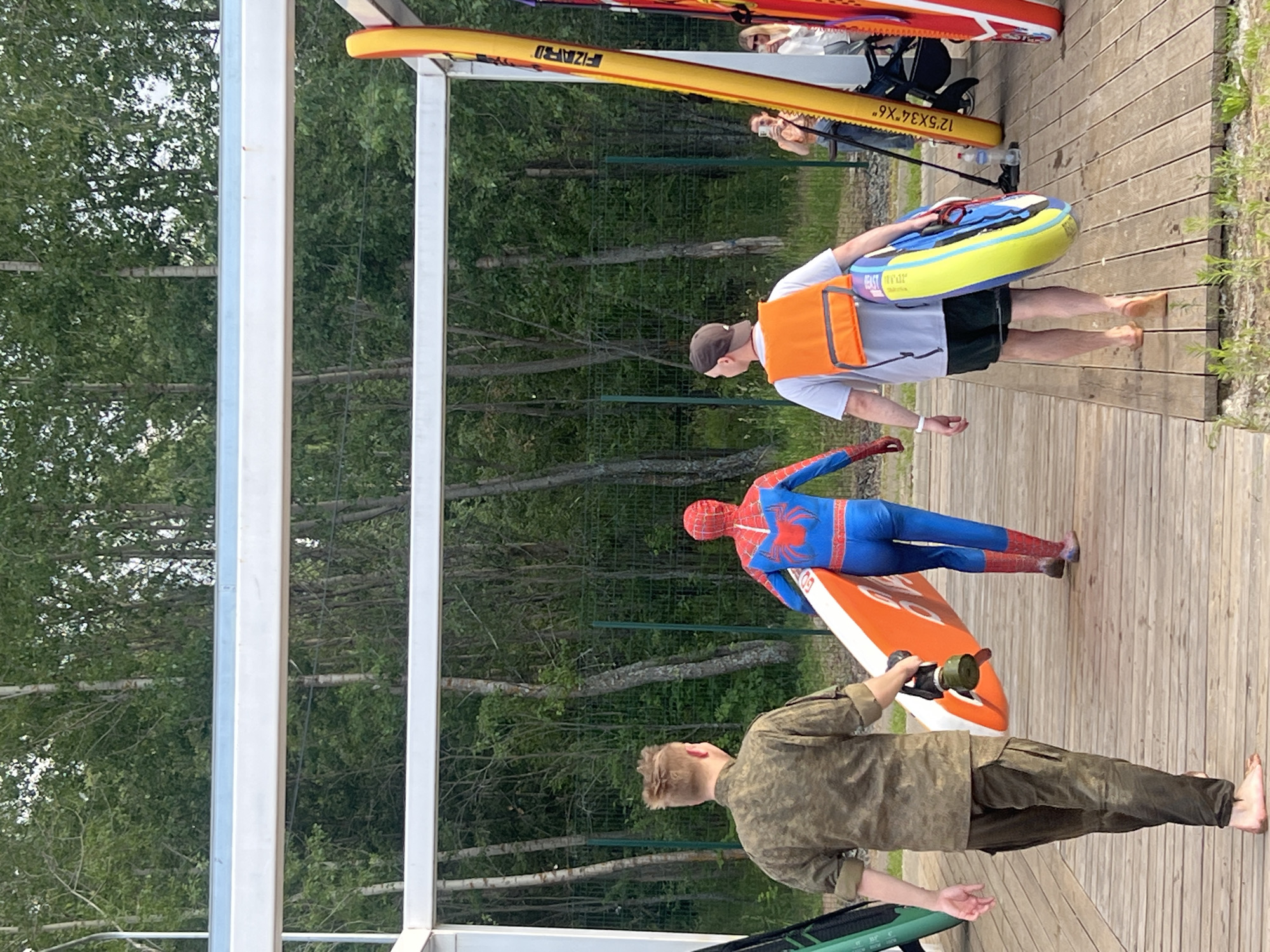 Three people are walking away from the camera on a wooden pier. The person on the left is wearing a camouflage uniform and carrying a round object attached to a paddleboard. The person in the middle is dressed as Spider-Man and is carrying an orange paddleboard. The person on the right is wearing a life vest over a t-shirt and shorts and is carrying a colorful inflatable tube. In the background, there is a dense forest and a metal fence. The scene appears to be at a recreational area near the Kama River in Chaikovsky, Russia.