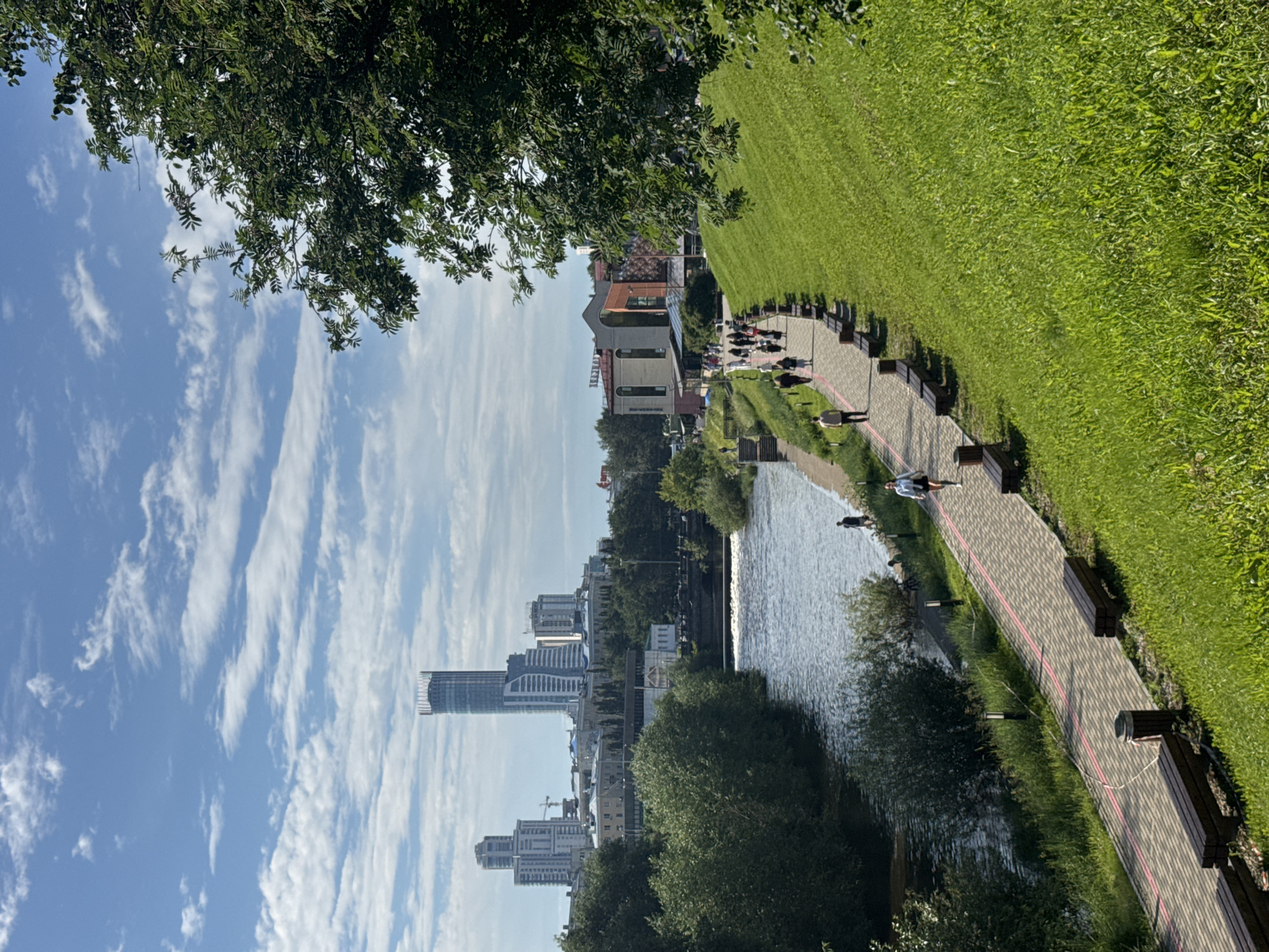 The image captures a lively scene in Yekaterinburg, Russia, on a clear, sunny day. The perspective is from a tree-lined area, looking down towards a river and a cityscape. The sky is a bright blue, dotted with wispy white clouds, suggesting pleasant weather.
In the foreground, lush green trees frame the top left of the image, their leaves dappled with sunlight. Below, a paved pathway runs alongside the river, bordered by manicured green grass and a row of dark wooden benches. Several people are visible on the pathway and near the riverbank. A person is jogging along the path, while others are walking or sitting. Two individuals can be seen closer to the water's edge.
The river itself is calm and reflects the sky and surrounding greenery. On the other side of the river, the Yekaterinburg skyline rises, featuring a modern skyscraper with a distinctive curved facade, as well as other mid-rise buildings and some construction cranes. The buildings appear to be a mix of residential and commercial structures.
The scene evokes a sense of an active and pleasant day in a city park. The presence of people enjoying the outdoors, the well-maintained greenery, and the juxtaposition of nature with the urban landscape create a feeling of vibrancy and relaxation. The time of day appears to be late morning or early afternoon, given the bright sunlight and the shadows cast by the trees and buildings. No visible text is present in the image.