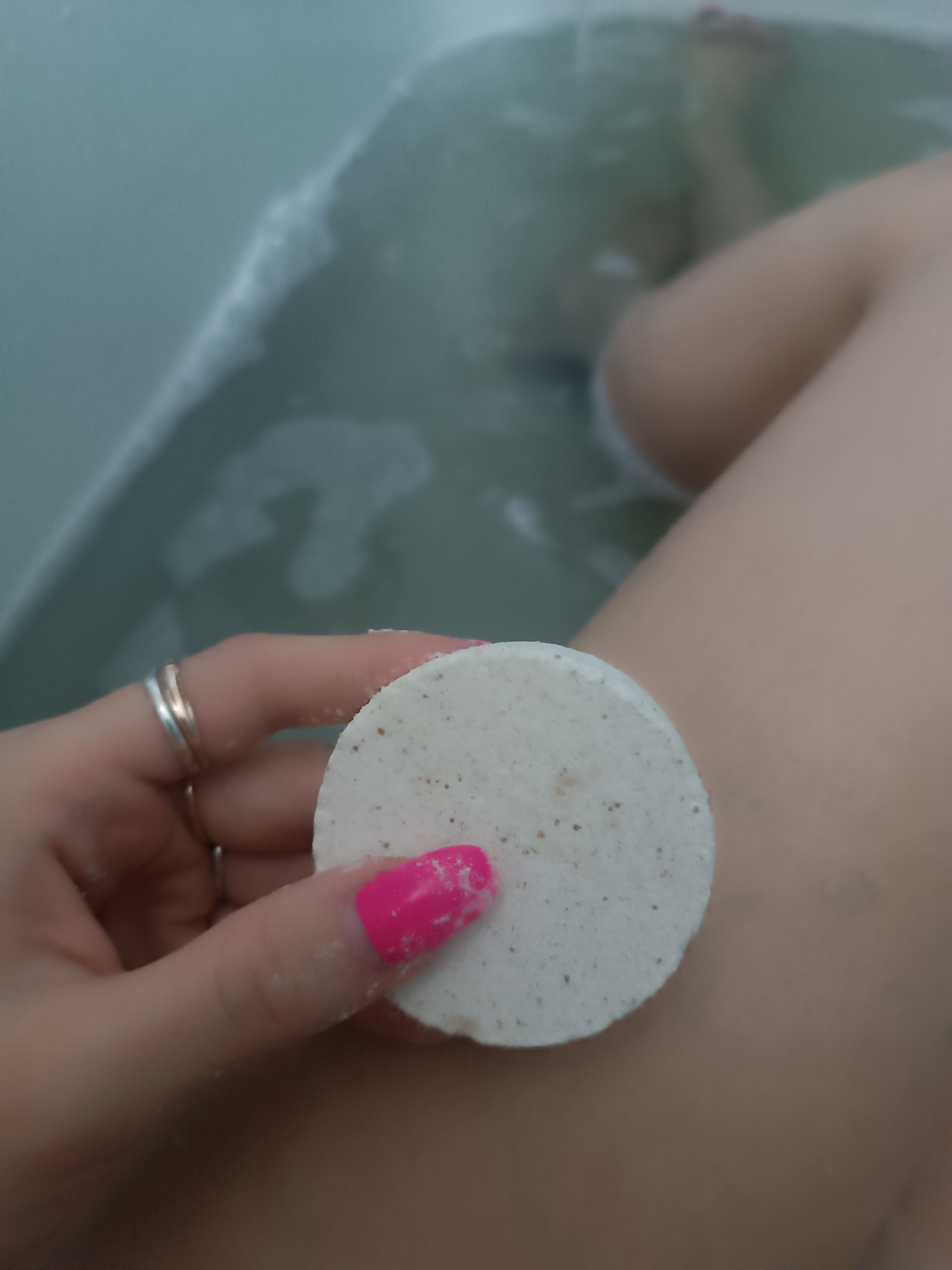 A close-up shot shows a hand holding a circular, speckled, off-white bath product, possibly a bath bomb or shower steamer. The hand has two silver rings on the ring and pinky fingers, and one fingernail is painted bright pink. The background shows a portion of a leg, implying the person is in a bathtub. Bubbles are visible in the bathwater, and the bathtub appears to be pale blue or grey. The location is unknown.
