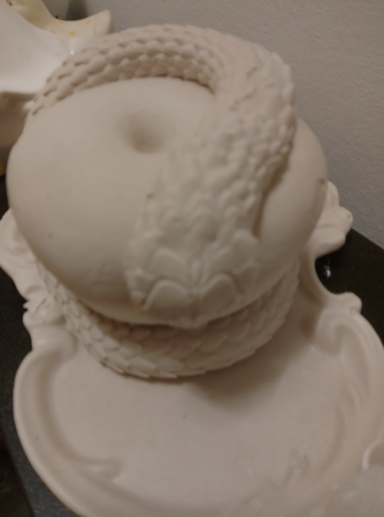 A white, ceramic, donut-shaped object is decorated with a raised, braided pattern around its upper surface. The object rests on a white, saucer-like base with a raised rim. The background is a plain, light-colored wall, and the surface beneath the object is dark. There is another white ceramic object to the left, partially visible and out of focus. The scene is a still life.