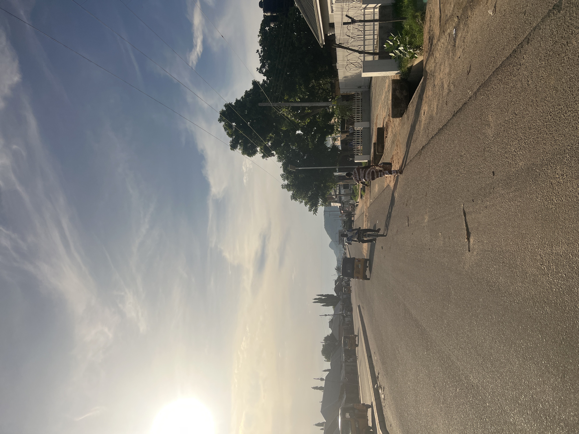 The image depicts a sunlit street scene in Jalingo, Nigeria, likely in the morning or late afternoon, given the warm, low-angled sunlight. A wide, two-lane asphalt road extends into the distance, appearing relatively clear of heavy traffic.
The sky is bright and expansive, displaying a pale blue hue with scattered, wispy white clouds. Power lines stretch across the upper portion of the sky, connecting to poles on the right side of the road. The sun, positioned low on the left, casts a strong, radiant glare, creating long shadows that stretch across the street and the sidewalk.
On the right side of the road, a paved sidewalk or unpaved verge runs alongside a series of buildings and lush green trees. The buildings are low-rise, with one prominent white structure featuring a corrugated metal roof and a fence topped with barbed wire. Large, dense trees provide ample shade along this side of the street.
Several people are visible in the scene. One individual wearing a striped top stands or walks on the right side of the road in the mid-ground. Further down the street, another person is seen near a vehicle. Multiple tricycles, commonly known as Keke Napep, are present on the road, some appearing to be parked, including a distinct yellow one.
In the far distance, a mountain or large hill range is discernible against the horizon, providing a natural backdrop to the urban setting. The overall atmosphere conveys a sense of daily life unfolding under a bright, open sky. No discernible text is visible in the image.