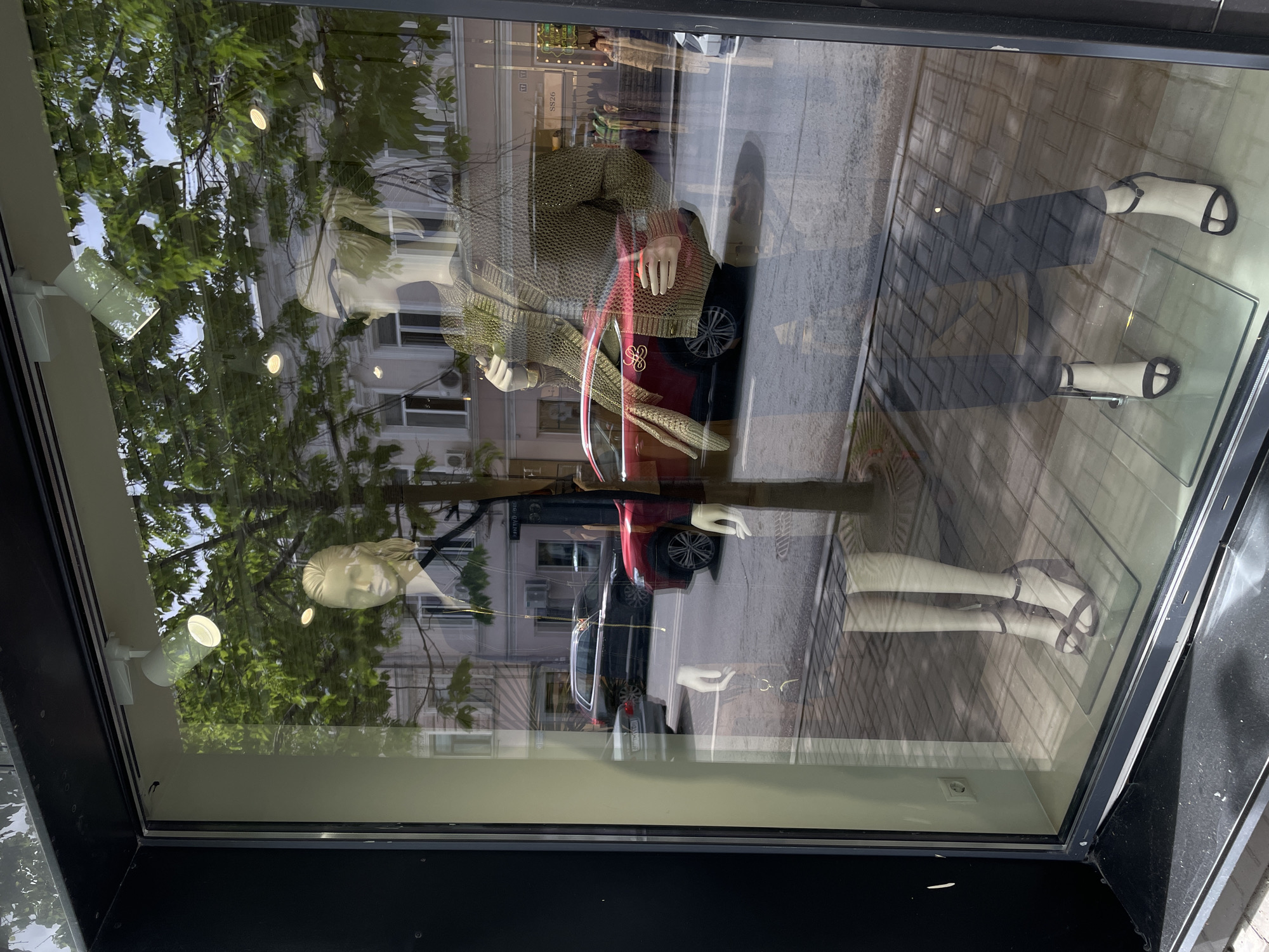 The image shows a storefront window display in Kazan, Russia. Inside the display, two mannequins are arranged: the top one wears a patterned sweater, a red bag, and black pants with white sandals. The bottom mannequin wears a long-sleeved red top and black pants with white sandals. The window reflection shows trees and a building, with cars parked on the street.