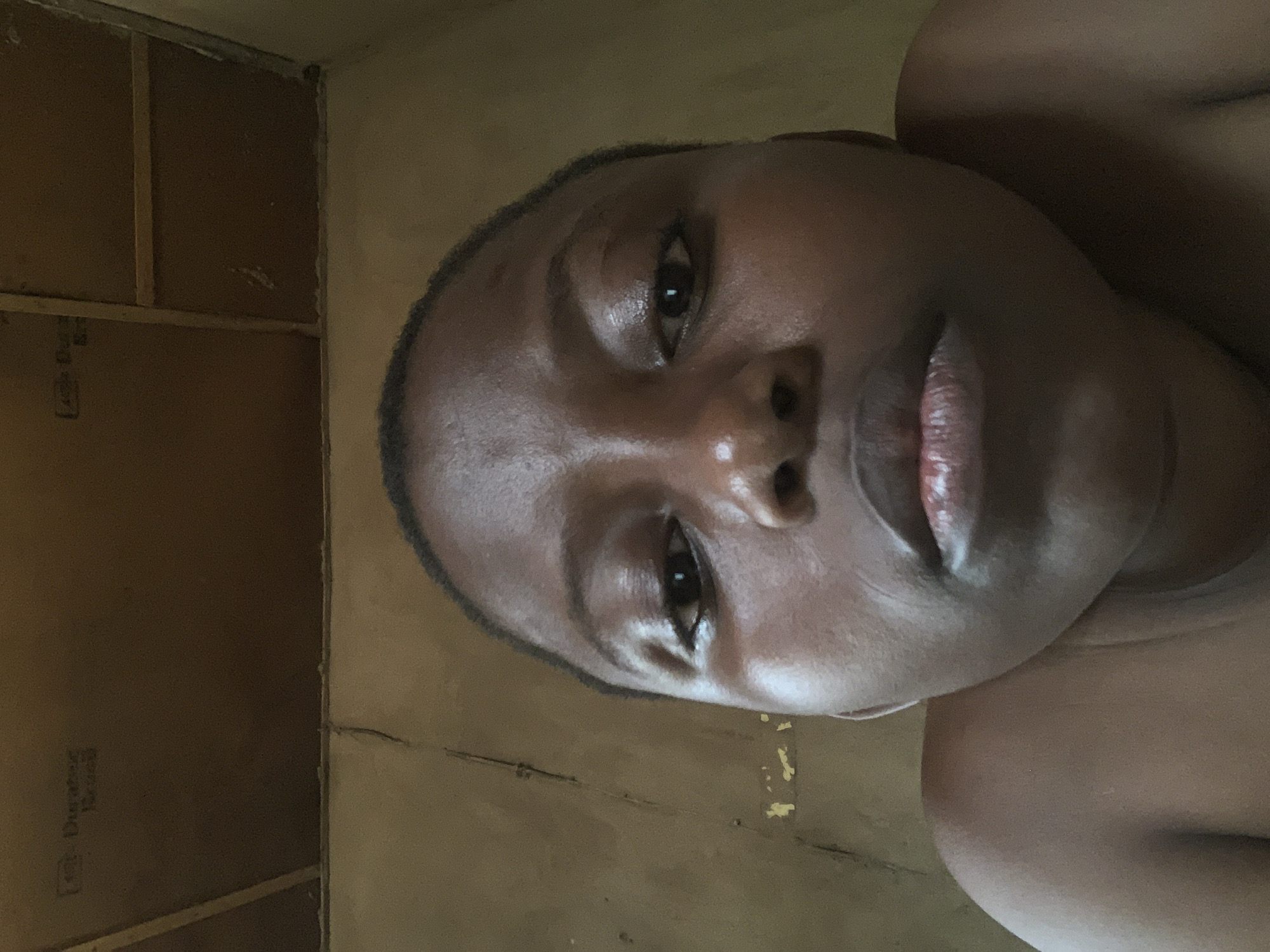 A close-up selfie of a dark-skinned person is captured. The individual has short hair and glossy skin. The setting appears to be indoors, with walls that look to be either under construction or in disrepair, potentially made of wallboards. The location, Ikorodu, Nigeria, cannot be confirmed based on the image content alone.