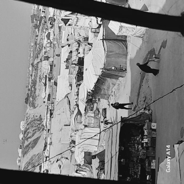 This black and white image presents a detailed view of a densely populated informal settlement or refugee camp located in Khan Yunis, Palestinian Territory, captured from an elevated position looking down into the camp. The scene is dominated by numerous makeshift tents and temporary structures, primarily constructed from light-colored tarpaulins and darker fabrics, tightly packed across undulating, dusty terrain.
In the foreground, on the left, an open-front stall or shop displays various goods on shelves and in crates, with a crescent moon symbol visible on one container. A dark diagonal cable or wire traverses the lower-mid section of the frame. On the uneven dirt paths, three individuals are present: on the right, an adult in dark clothing pushes a small, two-wheeled wooden cart or object, leaving a wet trail on the ground; another adult, also in dark clothing and wearing a cap, walks towards the mid-ground, looking down at a handheld device; and a child is seen running further into the camp.
The mid-ground and background are filled with more of these improvised shelters, extending up a prominent sandy hill or dune in the far distance. This hill is also dotted with scattered temporary structures and a few small, more permanent buildings at its crest. A tall communication tower or mast stands prominently on the highest point of the distant hill. The sky is bright and appears overcast. A "Galaxy A14" watermark is visible in the bottom-left corner of the image.