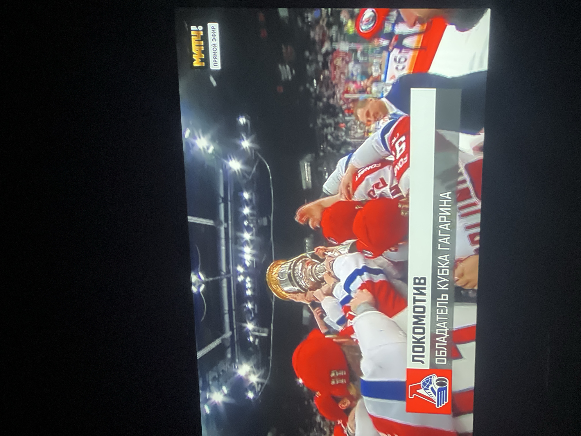 The image depicts a celebration scene, likely following a hockey game. Several people in red and white jerseys are gathered, lifting a golden trophy above their heads. A vertical banner displays the word "ЛОКОМОТИВ" and "ОБЛАДАТЕЛЬ КУБКА ГАГАРИНА" (Lokomotiv - Gagarin Cup Winners), indicating the team celebrating and the event. The background suggests a stadium setting with bright lighting. The "МАТЧ!" logo and "ПРЯМОЙ ЭФИР" (Live Broadcast) text are visible, confirming a television broadcast. There is no indication in the photo of Krasnoyarsk.