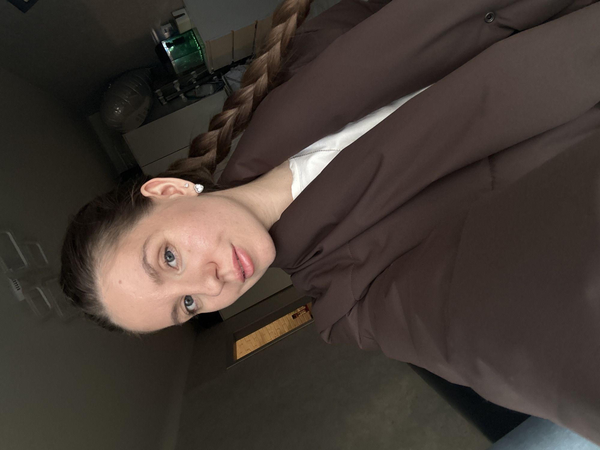 An image taken in what is stated to be a residential setting within the city of Klin, Russia, depicts a single female subject from the chest up, captured in a selfie. The woman, appearing to be in her 20s or 30s, has fair skin, light blue eyes, and light brown hair styled in a prominent braid extending over her right shoulder. She wears a brown puffer-style jacket over a white t-shirt with a thin silver chain necklace. Her left earlobe displays a heart-shaped earring adorned with small rhinestones, with a smaller stud visible above it. She looks directly at the camera with a neutral expression, lips slightly parted.
The background on the subject's right includes a white cabinet or shelf unit, upon which a small, illuminated aquarium containing green-tinted water and internal equipment is placed. Above this, a large, translucent, irregularly shaped object is partially visible. To the subject's left, a dark wall or furniture item is present, alongside an open doorway revealing a distant room with a light-colored brick wall pattern. Overhead, portions of a white ceiling and a rectangular light fixture are visible. The overall lighting is consistent with indoor artificial or subdued natural light.