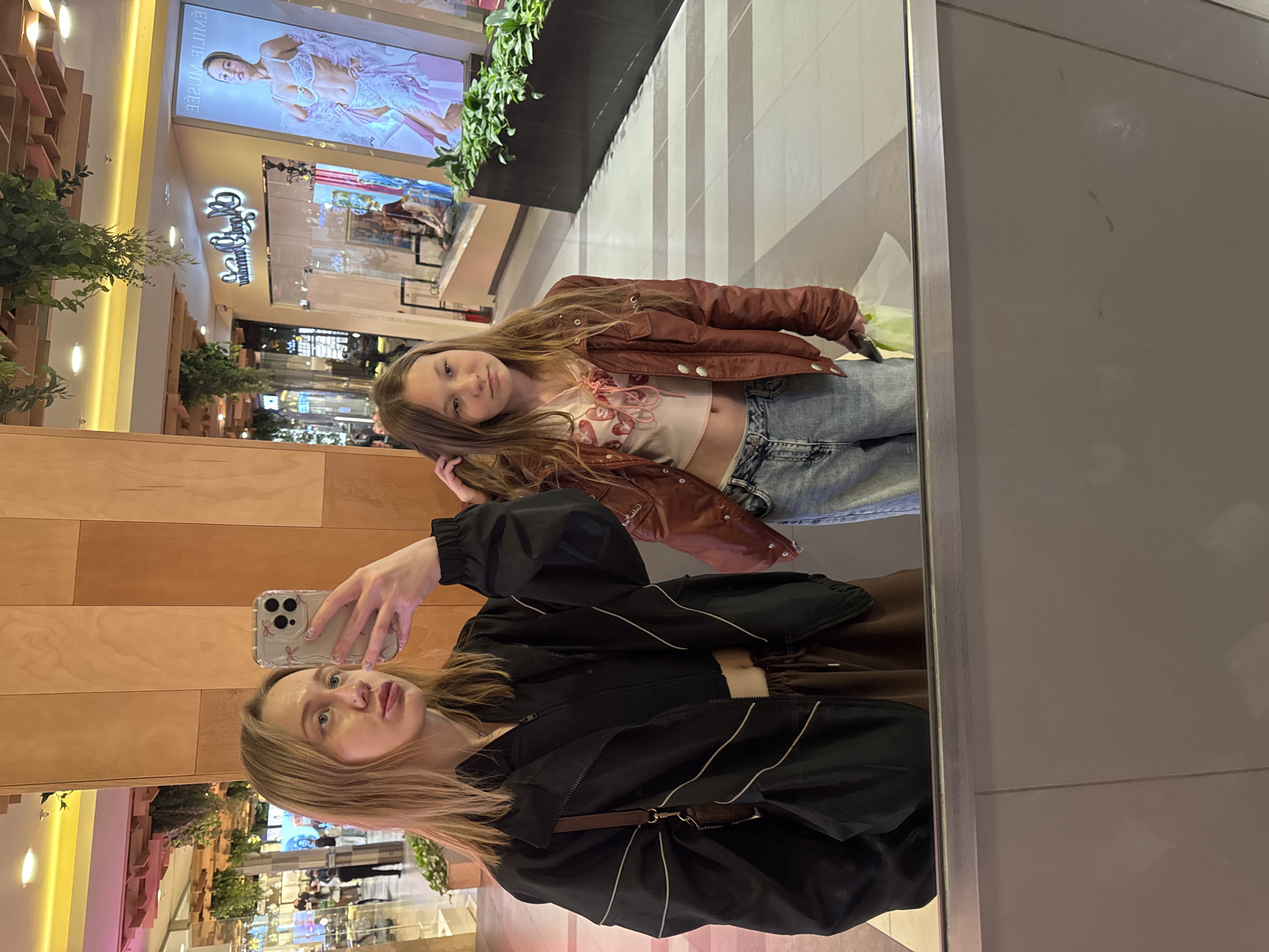 Two girls are taking a selfie in a mirror in a mall. One is holding the phone and wearing a black jacket. The other is standing behind her, wearing a brown jacket and jeans. The mall has multiple levels with stores and plants. The location is most likely Ol'ginskaya, Russia.