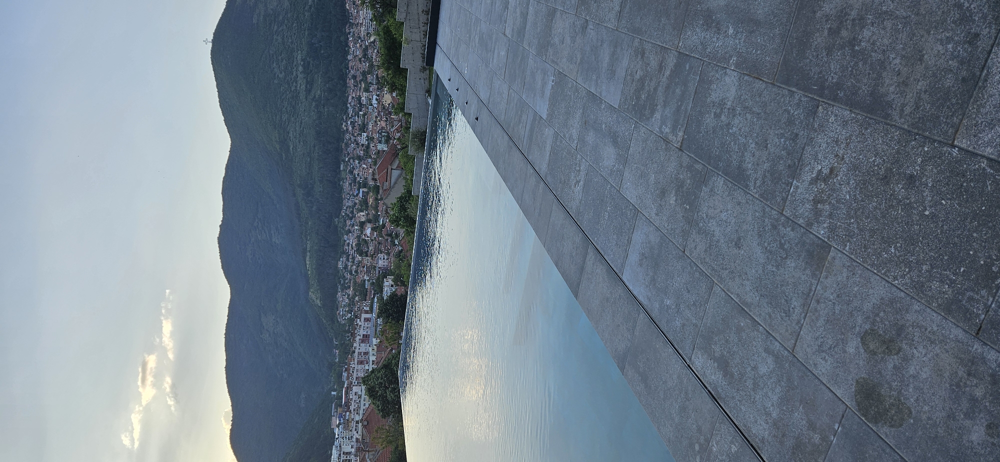A panoramic view showcases a hillside dotted with dense housing, adjacent to an infinity pool with a tiled edge. In the distant background, a mountain rises, marked by a cross atop its peak. The city is Plovdiv, Bulgaria. The pool's surface reflects the sky, creating a sense of seamless integration with the landscape. The scene is static, capturing a moment of tranquility.