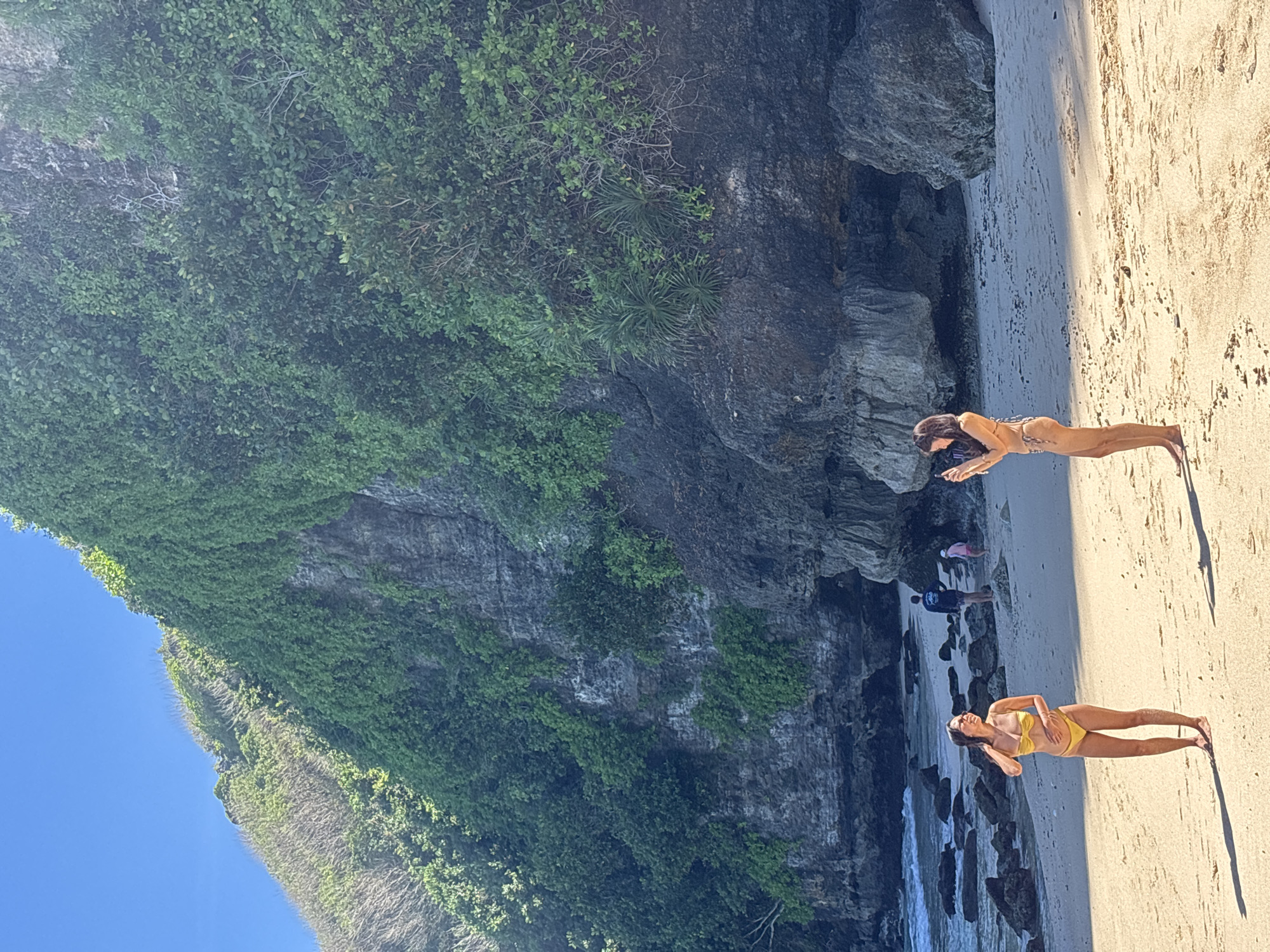 The image shows a beach in Kuta Selatan, Indonesia. On the right side of the image, there is a sandy beach, where two women are laying down. One woman is wearing a yellow bikini, while the other one is wearing a brown bikini. Behind the beach, there is a cliff covered in lush greenery. In the background, there are some people standing on the beach near the cliff. The sky is clear and blue.