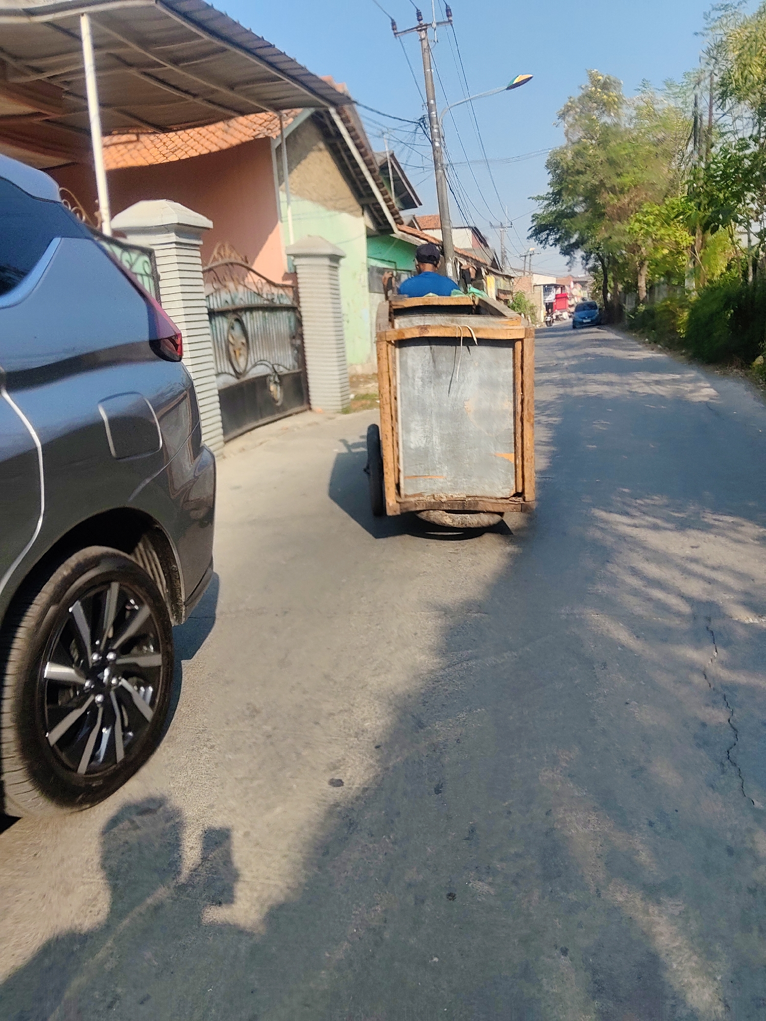 A person wearing a blue shirt and a dark cap is riding a three-wheeled cart on a street in Mekarmukti, Indonesia. The cart appears to be made of wood and metal and has a large rectangular container on the back. The street is paved and lined with trees and houses. A dark-colored car is parked on the left side of the road, and a shadow of what appears to be a person holding a phone is visible on the ground. The sky is clear and blue, indicating a sunny day.