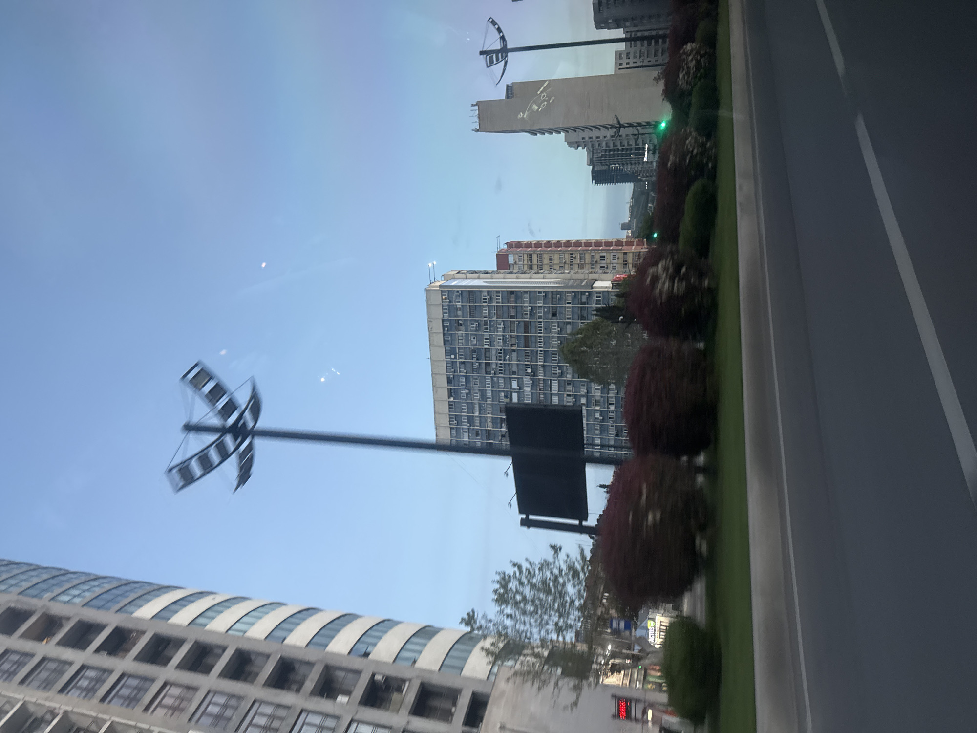 The image shows a street scene in Tbilisi, Georgia. A modern building with a distinctive curved facade and many windows is visible at the bottom. Above it is a tall, thin street light with a unique, abstract sculptural design at the top. Several high-rise buildings are scattered in the background, including a taller rectangular building with a facade divided by multiple windows, and another one that appears taller, beige, and narrow. A line of bushes and green spaces separates the buildings from a wide, gray road with white markings, suggesting a divided highway or main road. The sky is clear and blue.