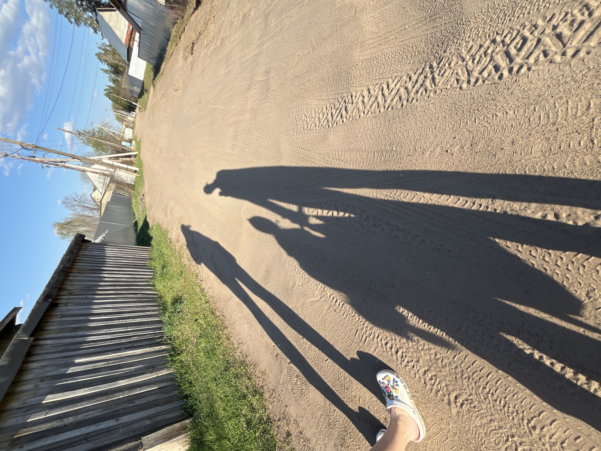 The photo depicts a dirt road in the city of Chaikovsky, Russia. The image is taken from a low angle, looking down the road. The shadow of the photographer and another person are visible in the middle of the road. A foot wearing a patterned Croc shoe is visible in the lower right corner. The road has tire tracks and is bordered on the left by a wooden fence and green grass. On either side are some houses. The sky is partly cloudy with blue patches.