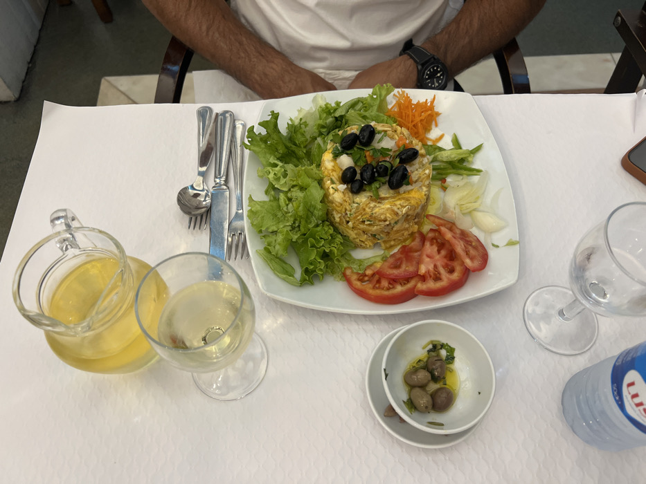 The scene depicts a restaurant table in Coruche, Portugal, covered with a white, textured tablecloth. A single individual's right arm is visible on the right side of the frame, wearing a white shirt and a black wristwatch.
Centrally placed on the table is a white rectangular plate containing a meal. The main component is a dish resembling shredded codfish with eggs and potatoes, garnished with whole black olives. This is surrounded by a fresh salad comprising green lettuce leaves, four slices of red tomato, shredded orange carrots, and thinly sliced white vegetables (likely onion or endive).
To the left of the main plate, two small white bowls are stacked; the top bowl holds green olives submerged in olive oil with visible herbs.
Beverages include a clear glass pitcher filled with a pale yellow liquid, presumably white wine, and a matching wine glass also containing the yellow liquid. Further down on the table, a plastic water bottle with a blue "LUS" branded label (likely Luso water) and an empty wine glass are visible.
A set of silver-colored cutlery, consisting of two forks, a knife, and a spoon, is neatly arranged on the tablecloth to the right of the filled wine glass. A dark brown object, possibly a phone or wallet, is partially visible near the empty wine glass.