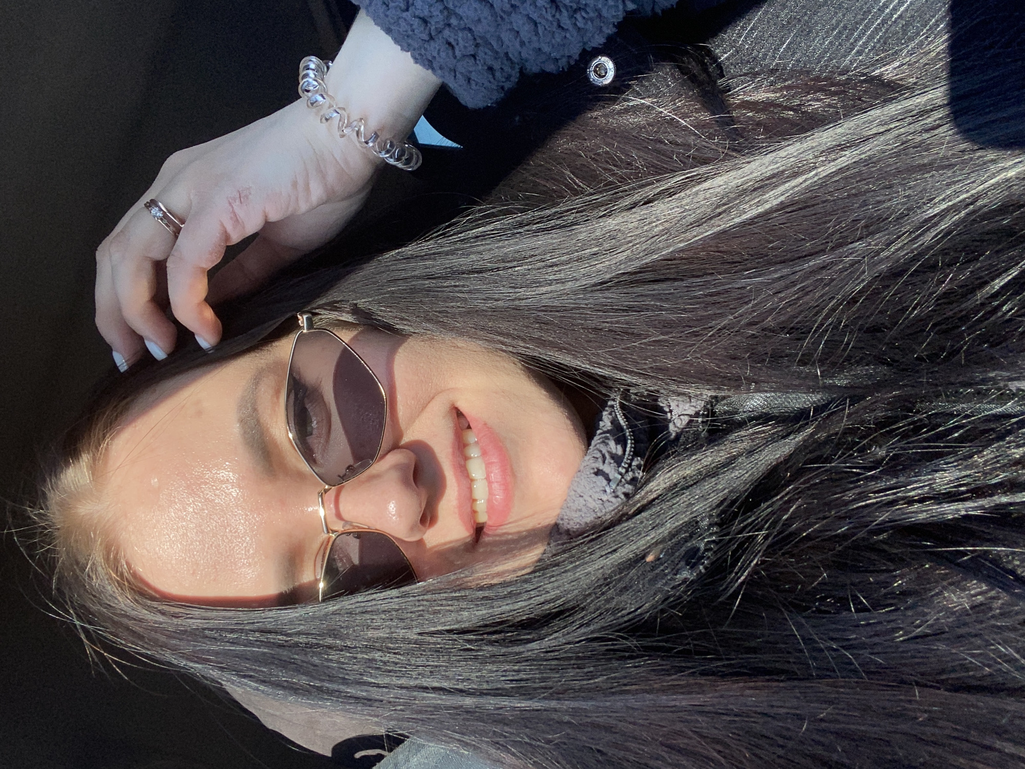 A woman lies on her side wearing sunglasses, a dark jacket, and a bracelet, smiling with her hand raised near her face. She is wearing a ring. The specific location in Votkinsk, Russia, is not identifiable from the image.