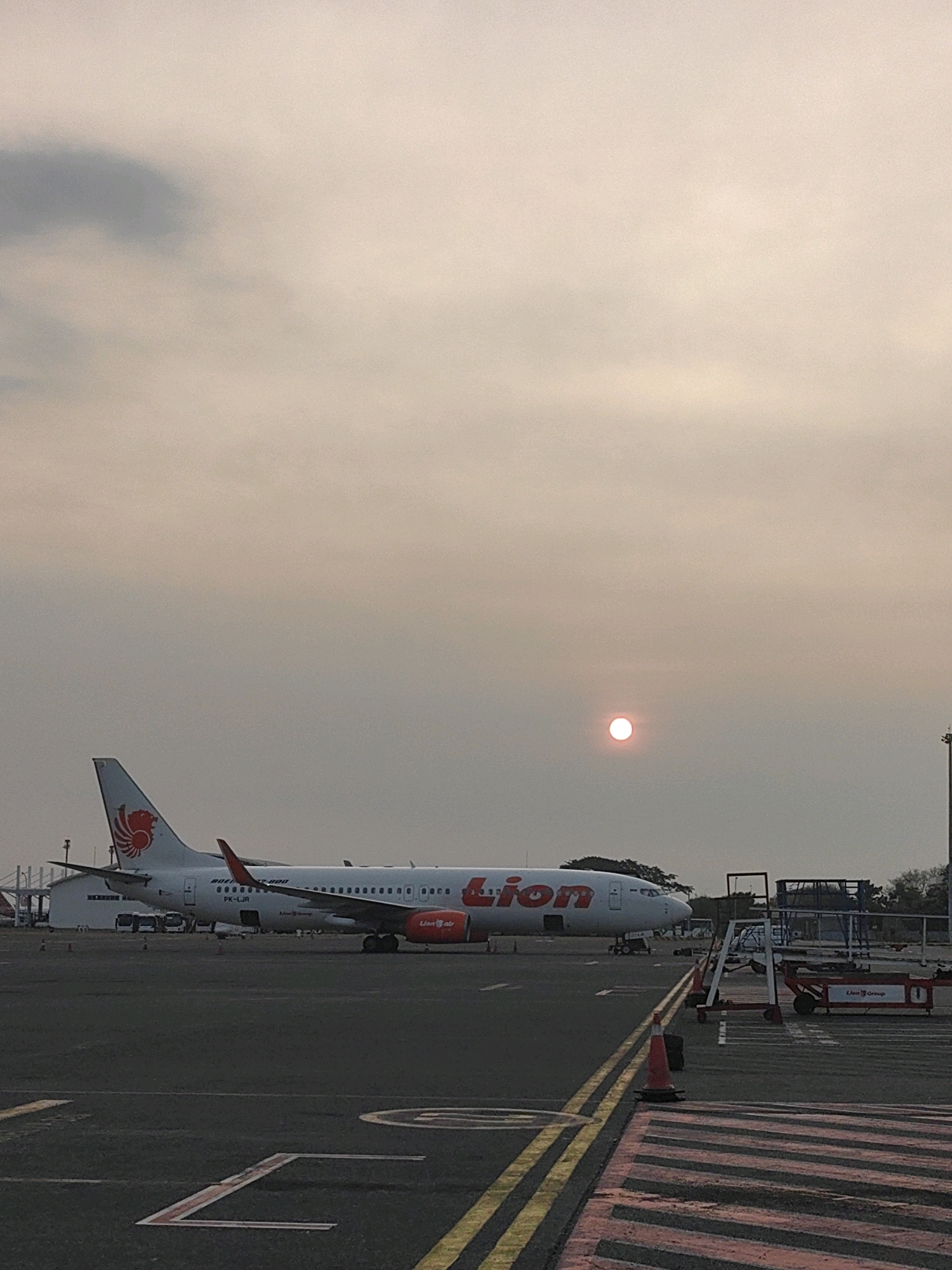An airplane with "Lion" written on its side sits on an tarmac in Segoro Tambak, Indonesia. The plane is white with a red and orange logo on its tail. The sun is visible in the sky, appearing as a bright orange orb behind a hazy sky. The tarmac is marked with yellow and red lines. In the background, there are buildings and trees. There are also some ground vehicles and equipment visible on the tarmac. The scene appears to be set during either sunrise or sunset.
