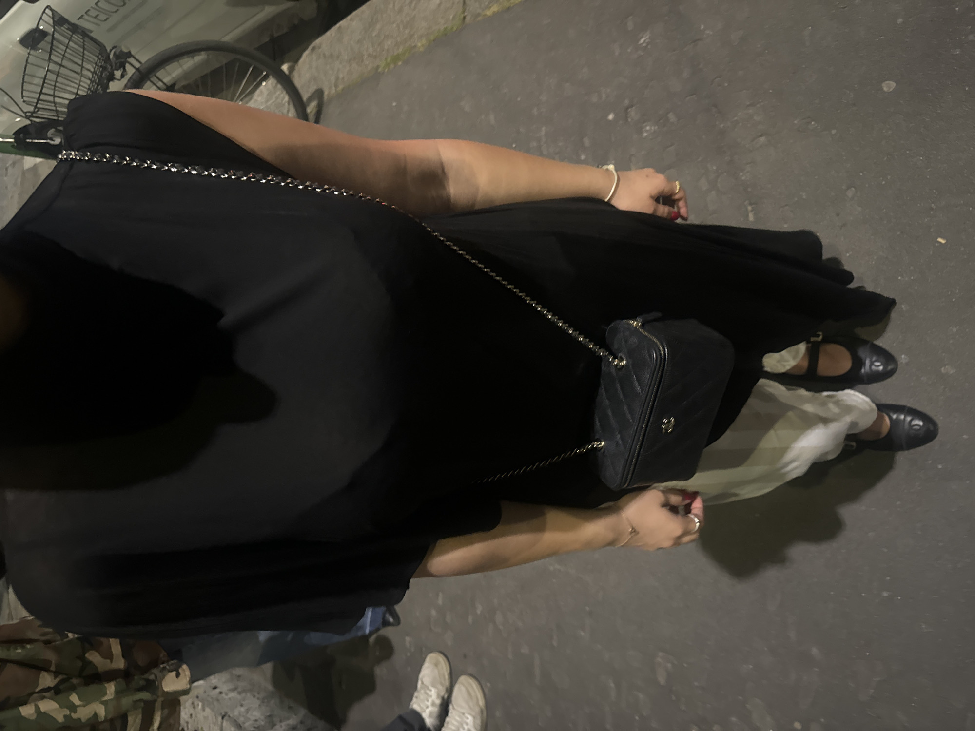 A medium shot captures a person in a dark dress with a quilted handbag, standing on a city street. The person has one hand and part of one arm visible, adorned with a gold bracelet. The hand has rings on two fingers and red nail polish on one. The handbag is suspended from a chain strap that crosses the person's body. The individual is wearing black shoes, and part of white pants is visible.
To the left of the person, a bicycle is parked. To the right, the street surface shows signs of wear and use. Below the person, there's a pair of sneakers and part of camouflage pants.