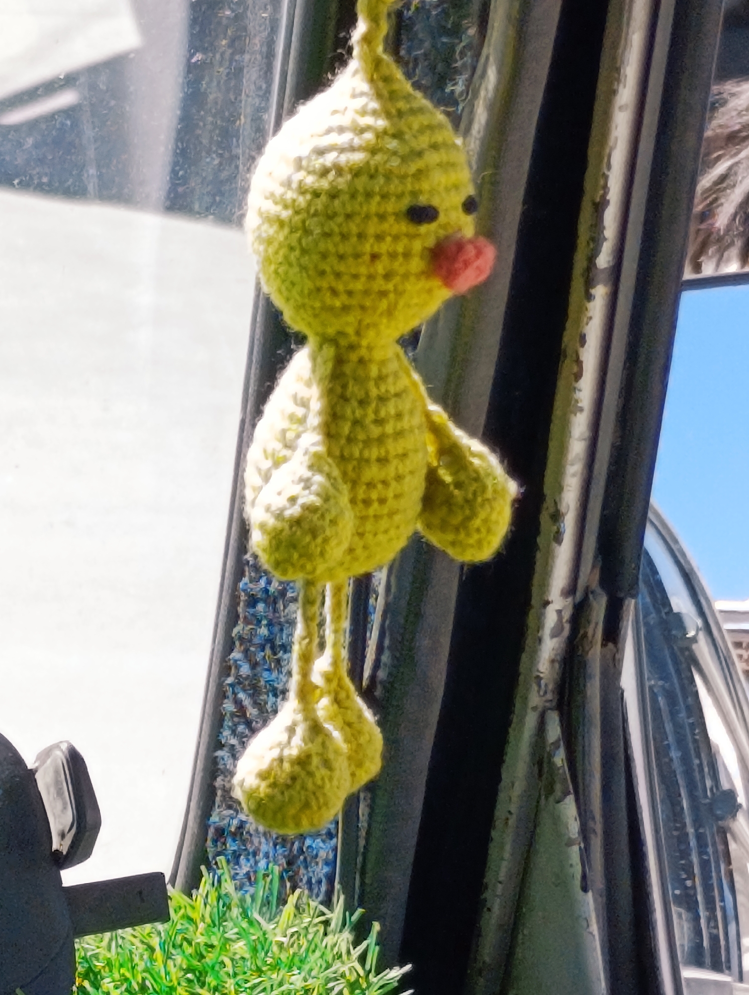 A bright yellow, crocheted chick hangs from what appears to be the interior of a vehicle, possibly a car. The chick is suspended by a string and has black eyes and a pink beak. It is positioned centrally in the frame, with a blurred background that suggests an outdoor environment. To the left, a black car door handle and a portion of the car's interior are visible. To the right, the dark frame of the car door is prominent, with glimpses of a bright blue sky and some foliage below. The image is taken in the city of Leh, India.