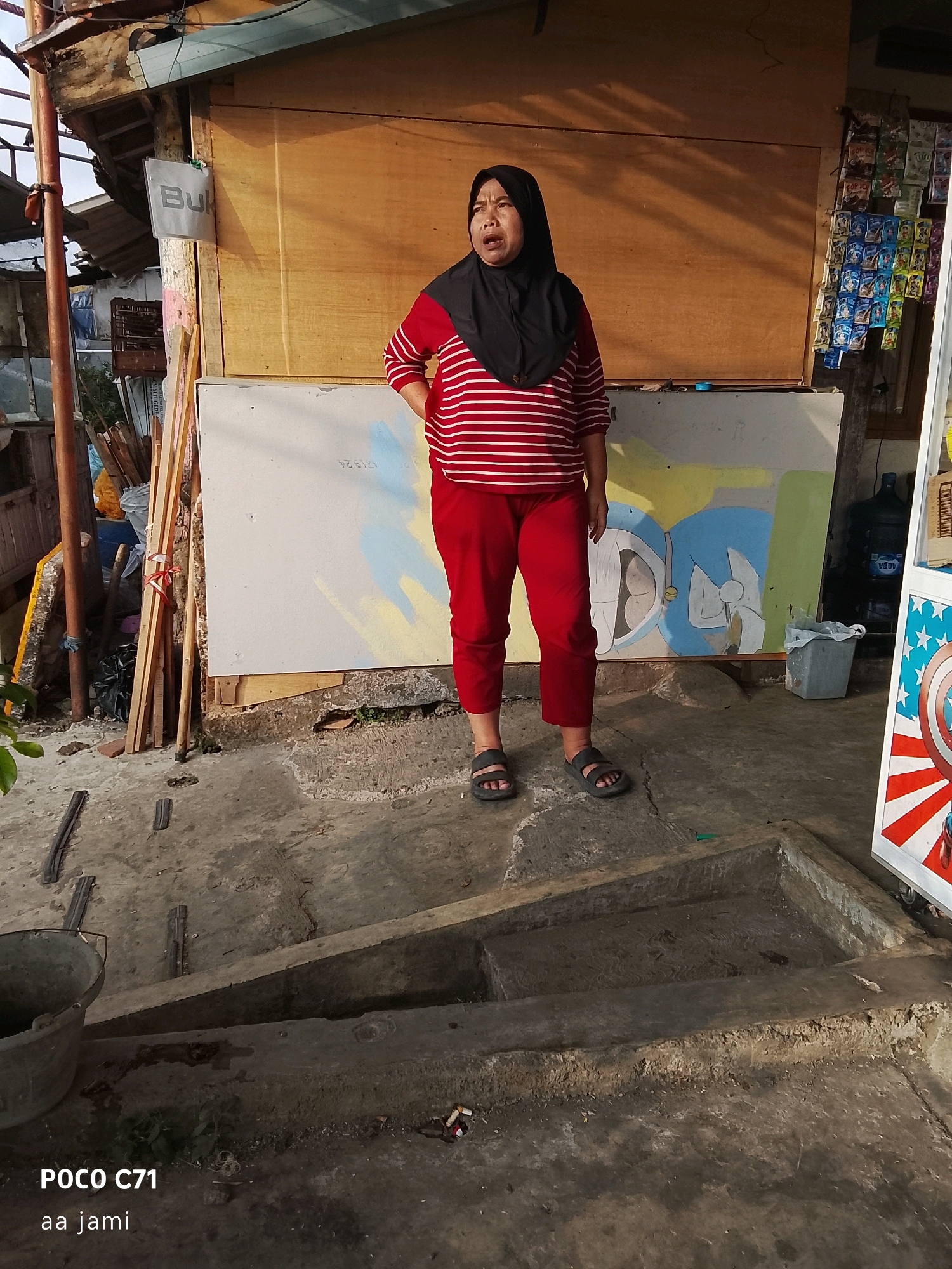 A woman wearing a red and white striped shirt, red pants, and a black hijab stands in front of a wooden stall in Tasikmalaya, Indonesia. She is looking upwards with her mouth open. Behind her, a white board with a cartoon drawing is leaning against the stall. Various items, including bags of snacks and a large water jug, are visible on shelves to the right of the woman. The ground is concrete and appears worn. In the foreground, a concrete structure resembling a trough is present.