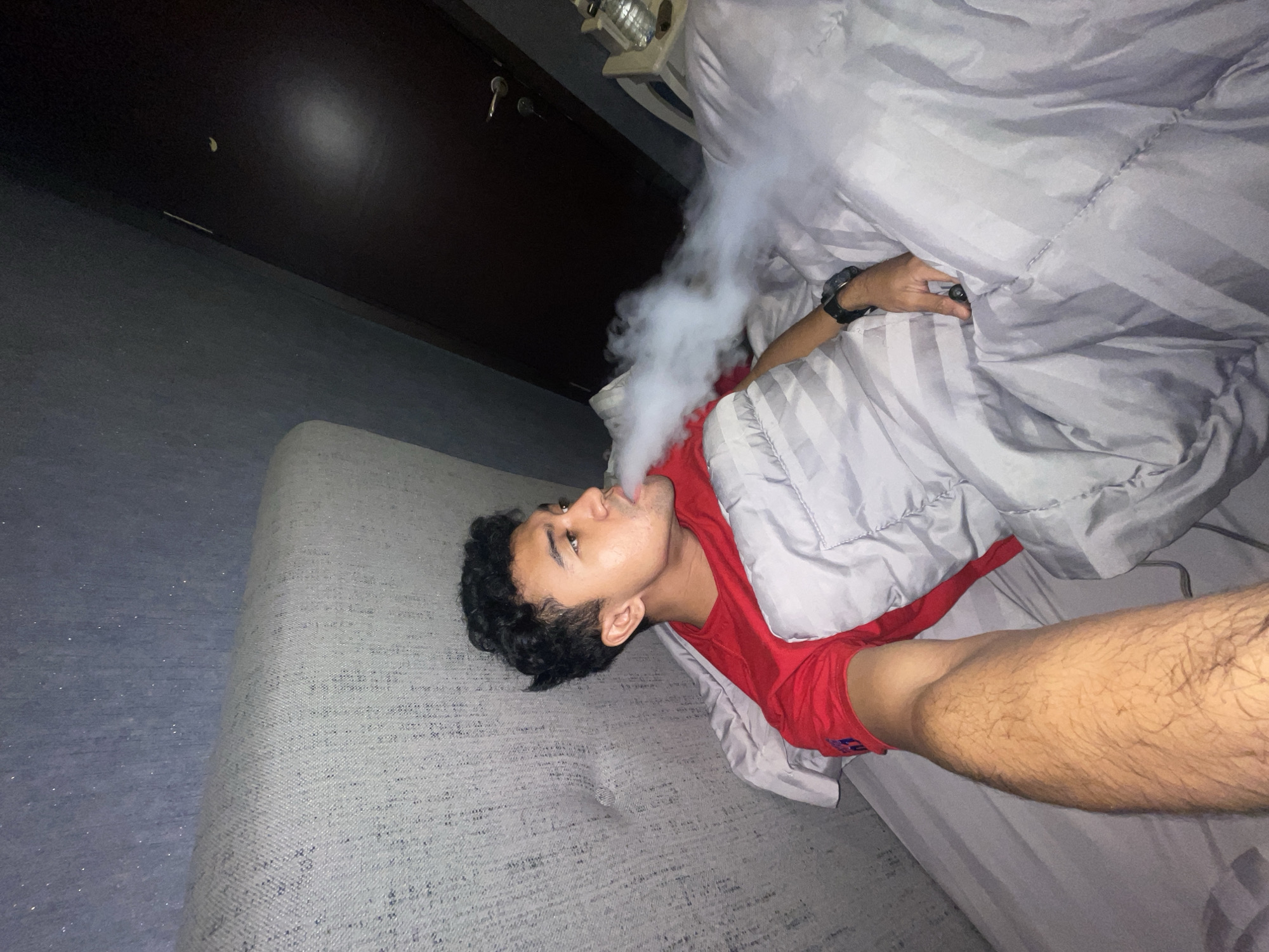 A young man lies on a gray bed with a gray duvet, exhaling a large plume of vapor. He wears a red t-shirt and a black wristwatch. The scene is inside, with a dark-colored cabinet and a heater-like object in the background. No verifiable information places the location in Central Jakarta, Indonesia.