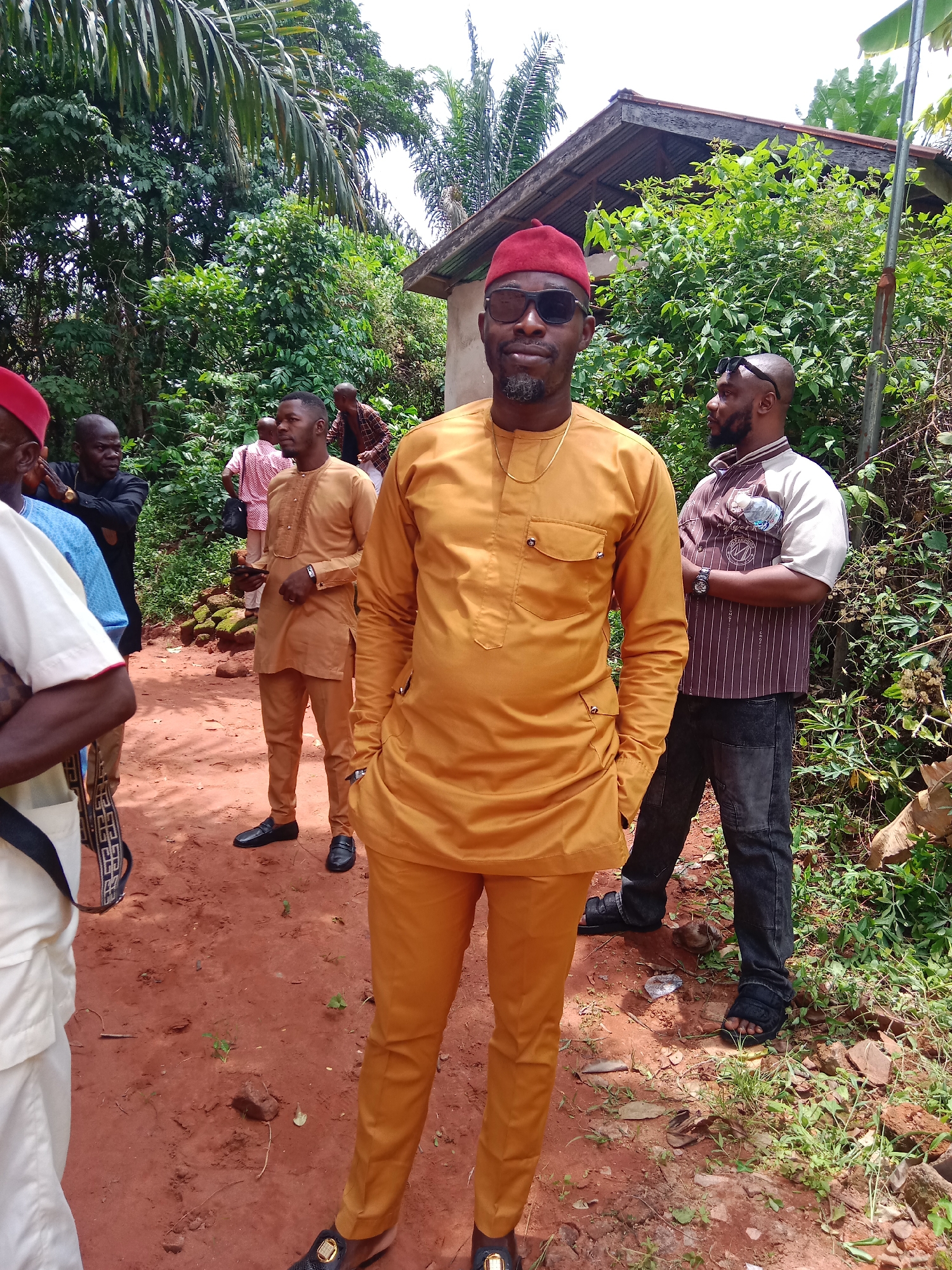 The image shows a group of men standing outdoors in Udi, Nigeria. The foreground features a man in a mustard-yellow traditional attire, complete with matching pants, a red hat, and dark sunglasses. He has a short beard and mustache. He is positioned with his hands in his pockets. Behind him and to the sides are several other men, some in similar traditional clothing but different colors and styles. The background consists of a dirt path surrounded by dense green foliage, trees, and a small structure with a rusty roof. The ground is reddish-brown dirt. The sky is overcast. The environment suggests a rural or semi-rural setting.
